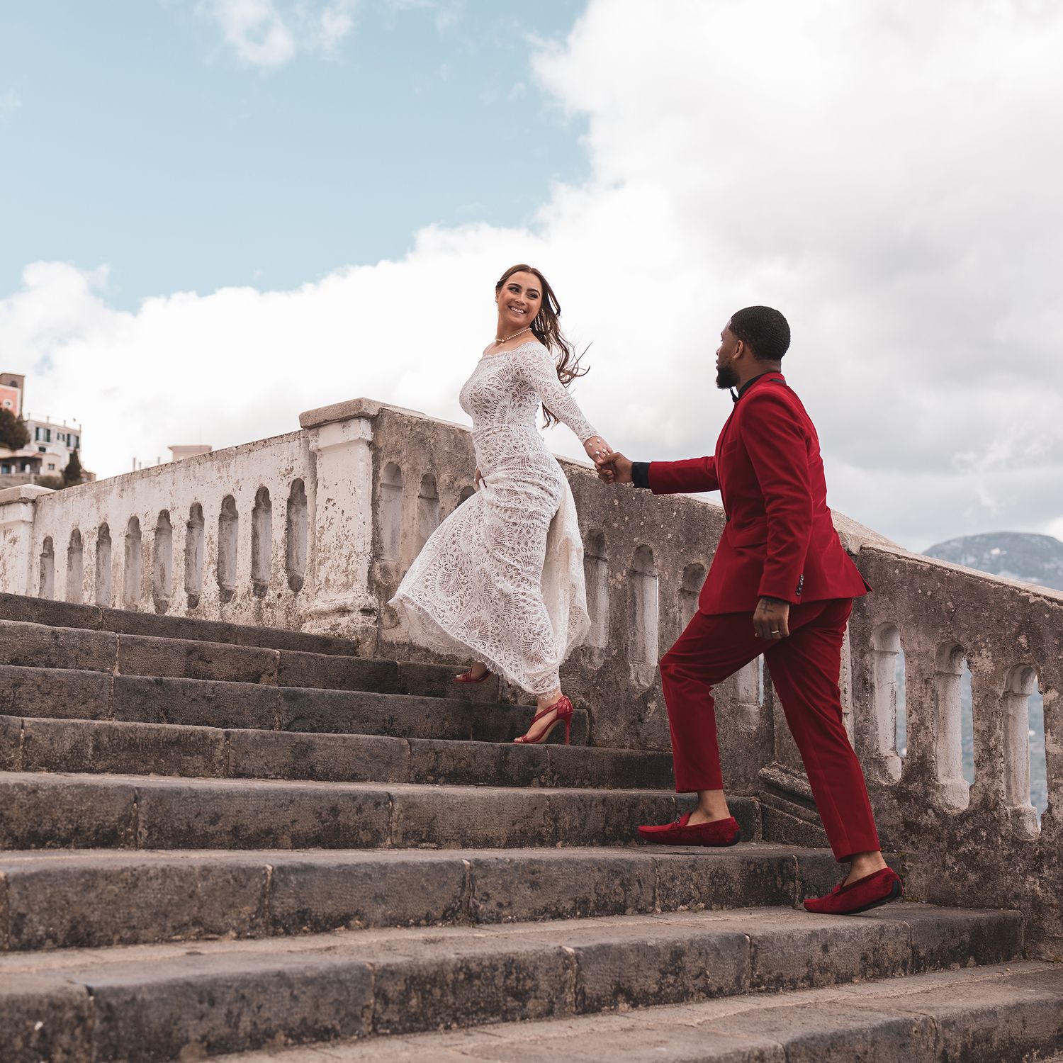 Couple in white dress and red suit holding hands while ascending stone steps at a historic building with mountain backdrop.