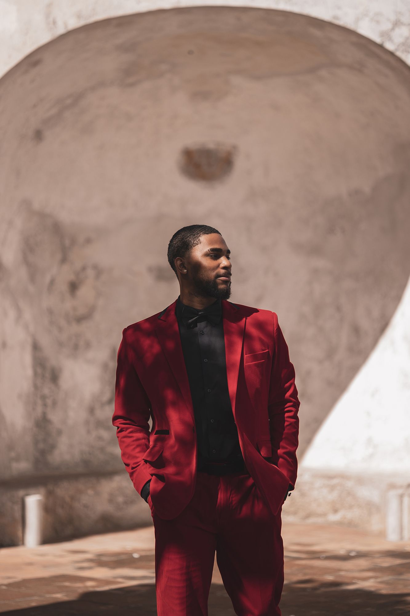 A person wearing a stylish burgundy red suit and black shirt poses against a curved concrete architectural backdrop.