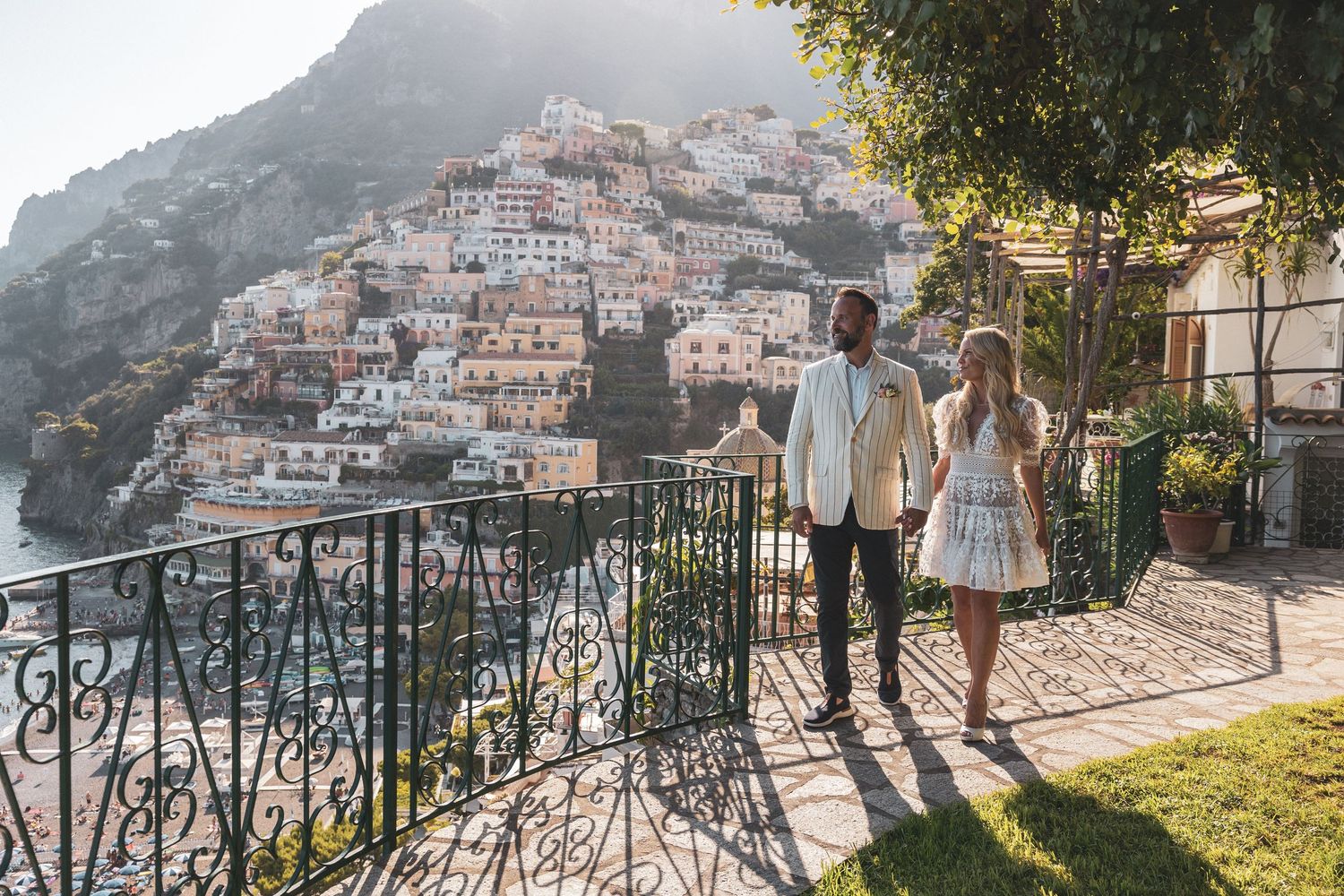 People stroll along a scenic overlook in Positano, Italy with white buildings cascading down the hillside in golden light.