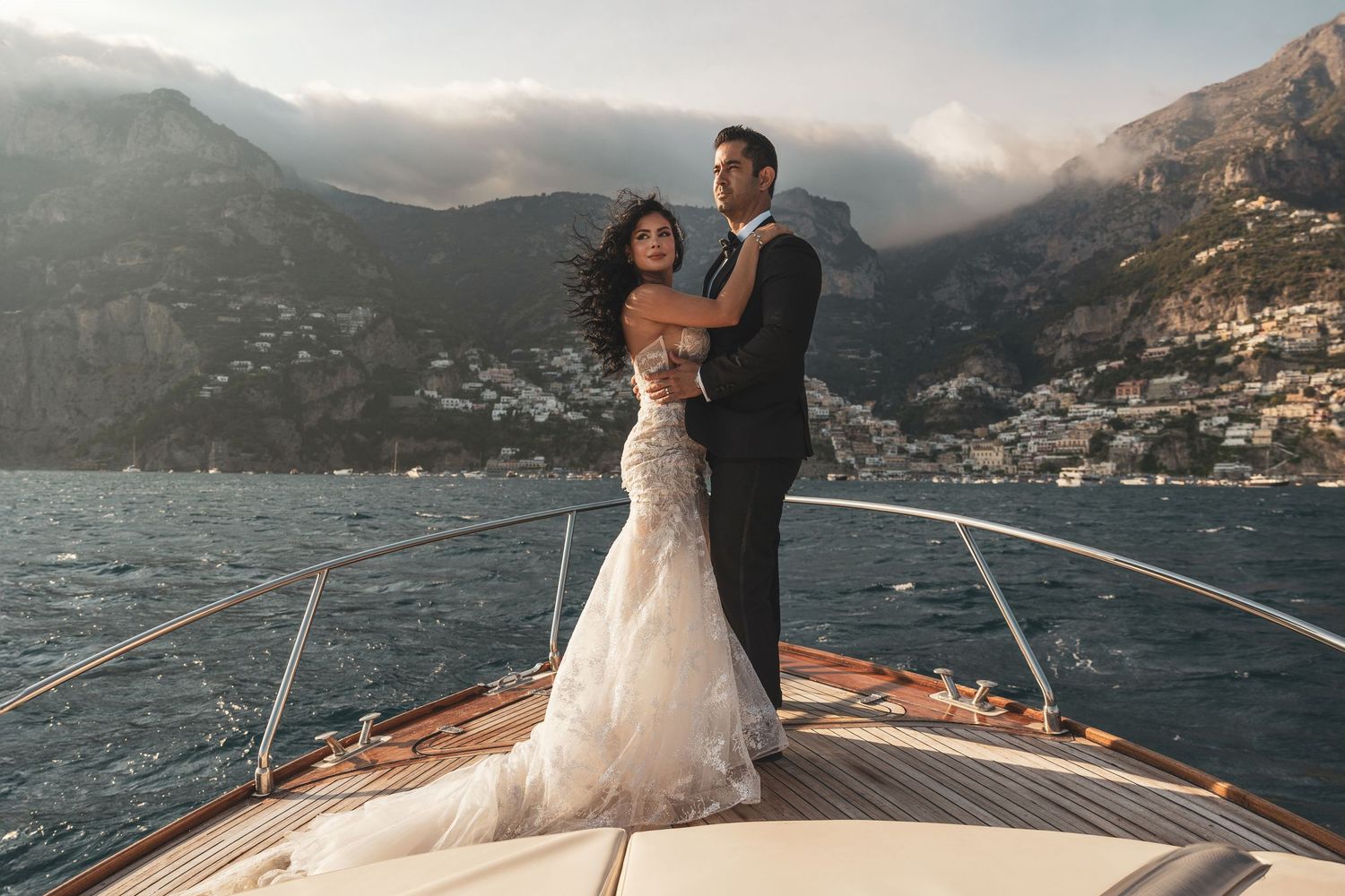 Romantic wedding couple embracing on luxury yacht deck with scenic coastal mountains in background at sunset.