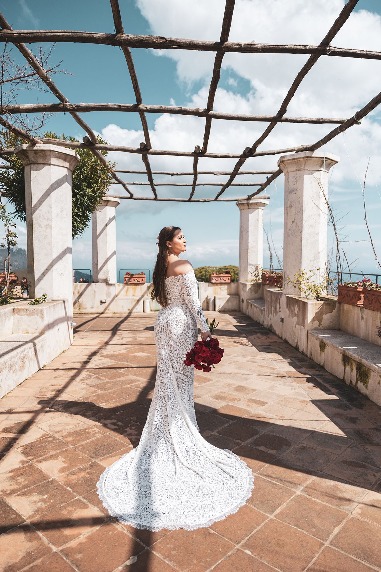 A bride in a form-fitting lace wedding gown holds a red bouquet under a Mediterranean pergola with ocean views.