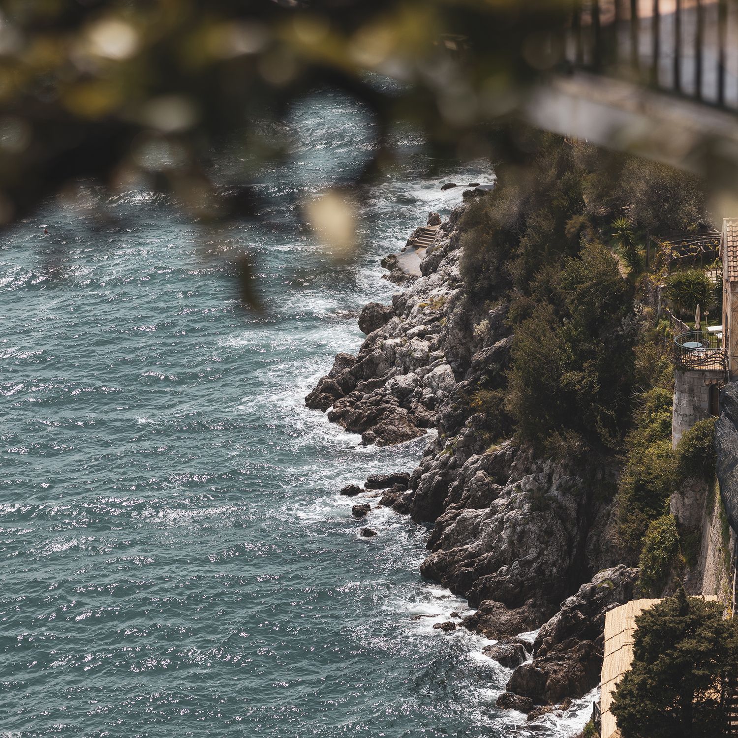 Aerial view of rocky coastline with turquoise waters crashing against steep cliffs on a misty Mediterranean shore.