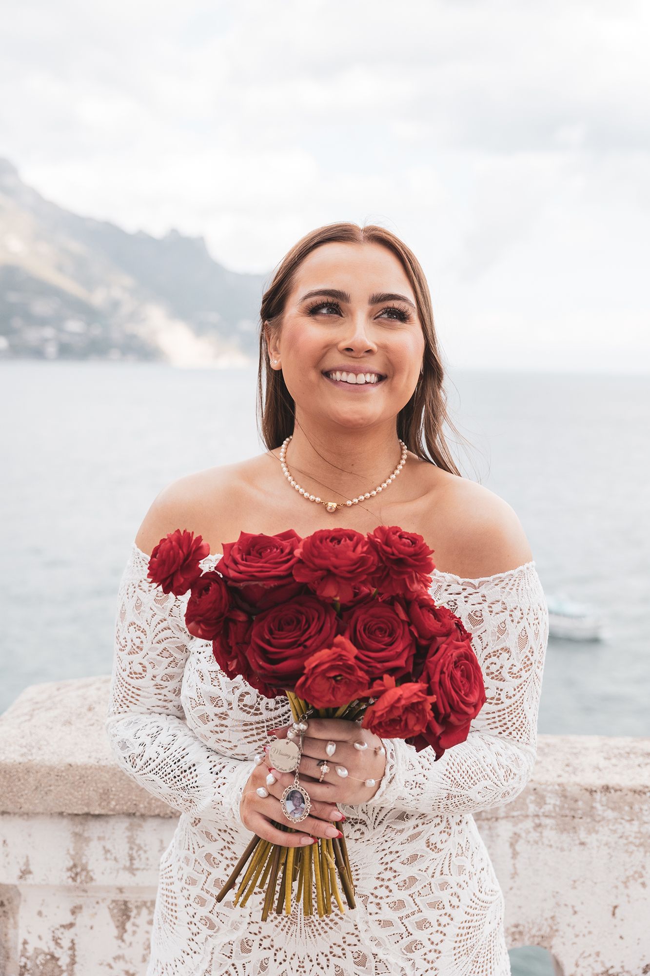 Bride in lace off-shoulder wedding dress holds vibrant red rose bouquet against coastal mountain backdrop.