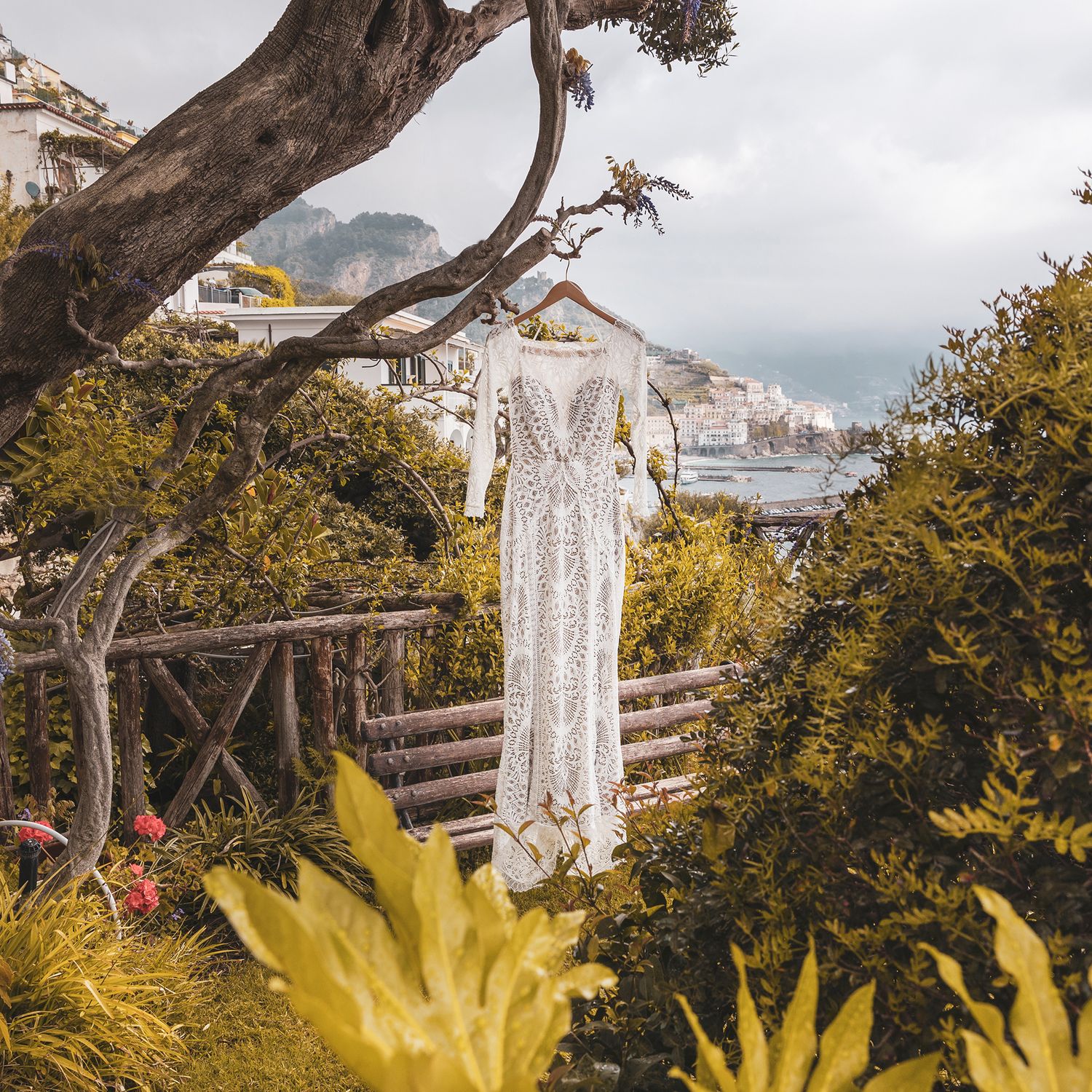 White lace wedding dress hanging on display in a garden overlooking coastal Italian town with mountains in background.