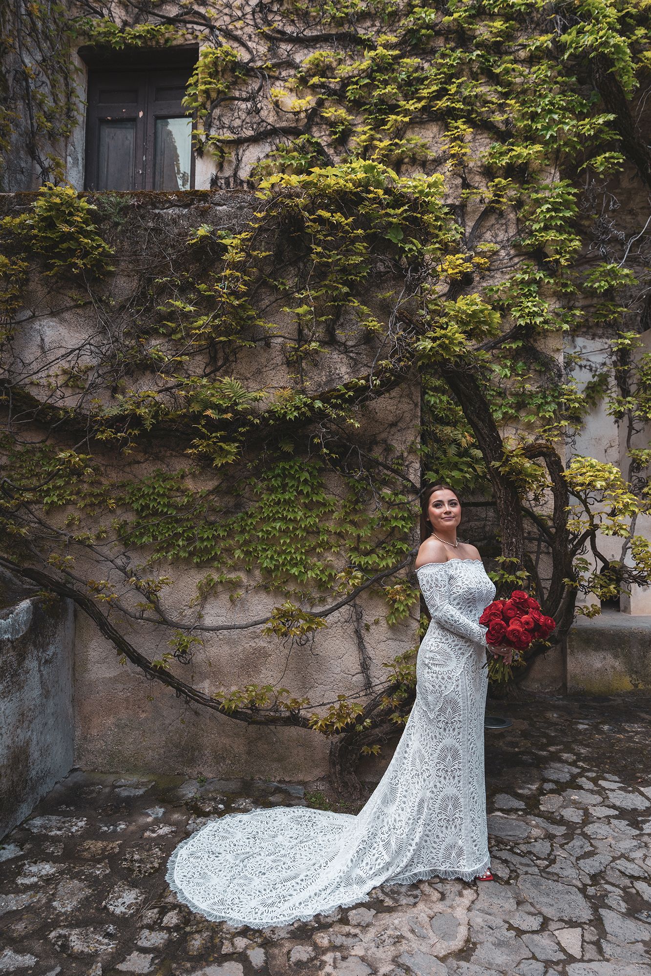 An elegant lace wedding dress with a long train contrasts beautifully against a vine-covered stone wall and cobblestone ground.