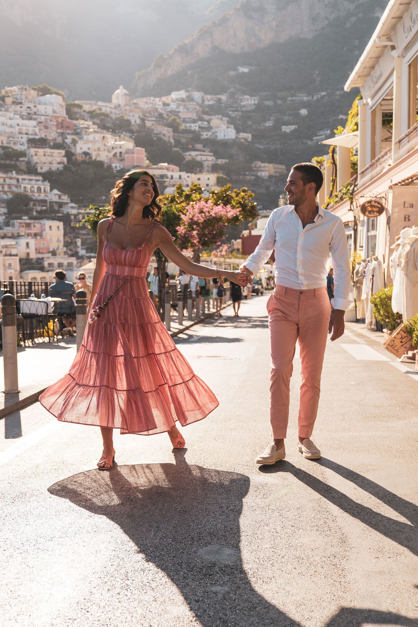 Two people walking down a sunny street in Positano, Italy with pink dress and white shirt against mountainside backdrop.