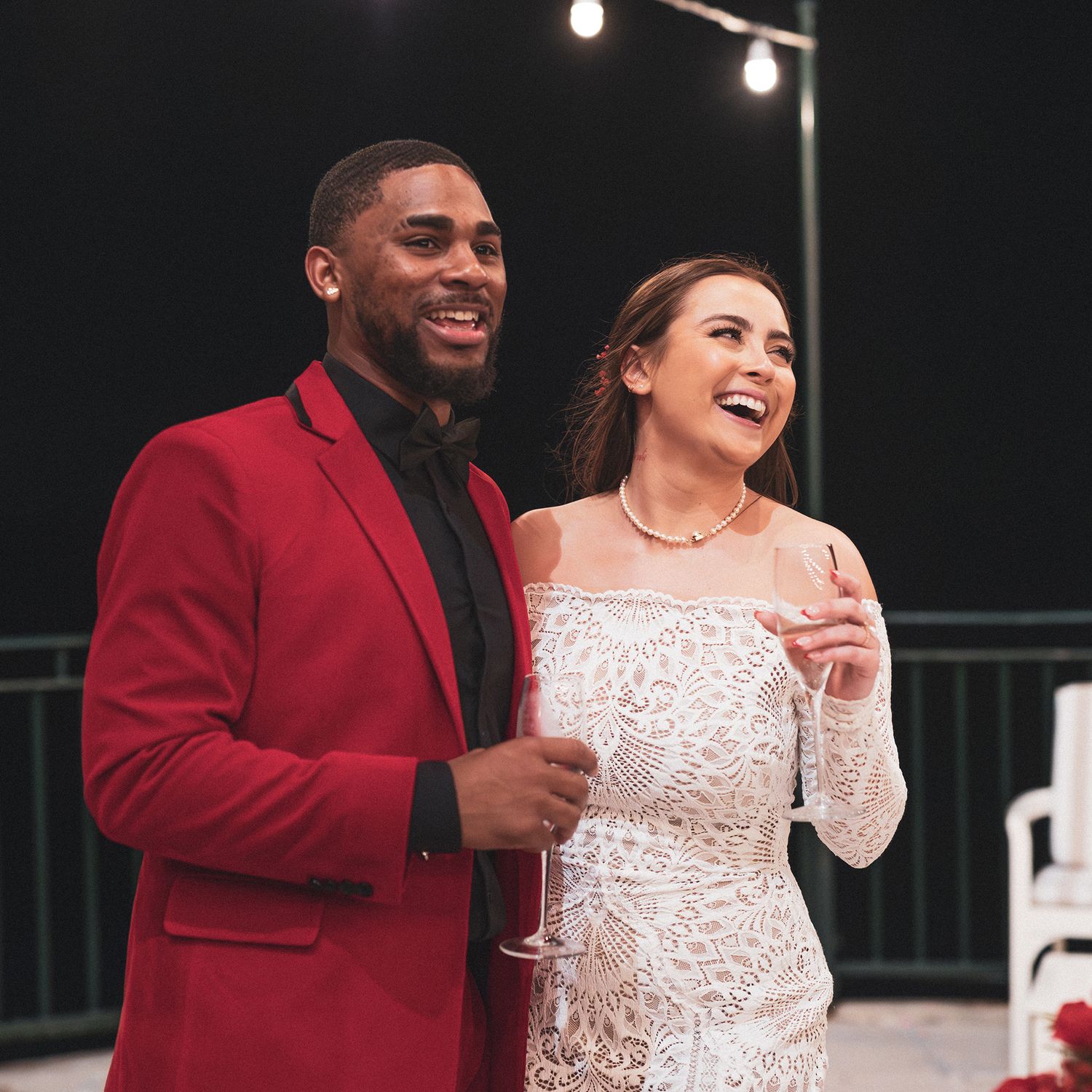 A couple laughs together at an evening celebration, with the groom in a red blazer and bride in a lace white dress.