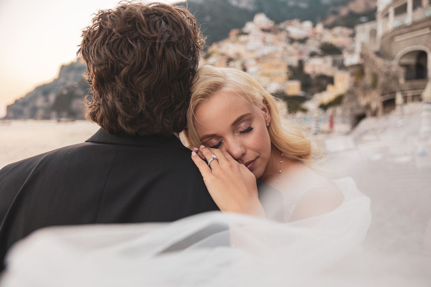 Romantic couple embraces on a beach during sunset with mountains and buildings in the background.