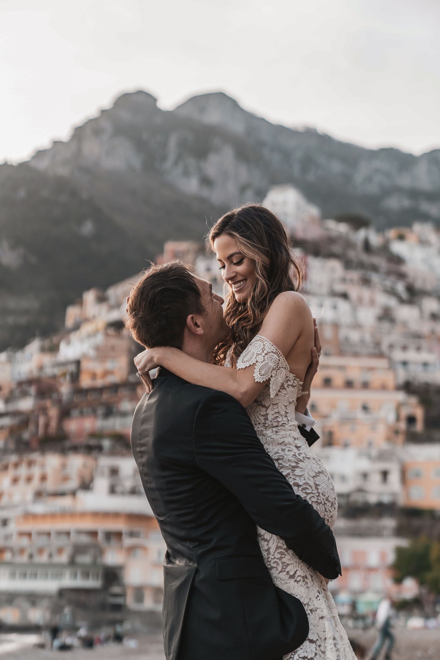 A romantic couple embraces against the scenic backdrop of Positano, Italy's colorful hillside buildings at sunset.