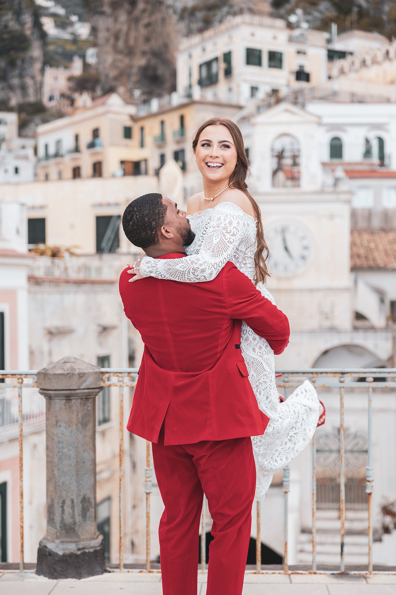 A joyful couple embraces on a scenic balcony overlooking the cliffside buildings of Positano, Italy.