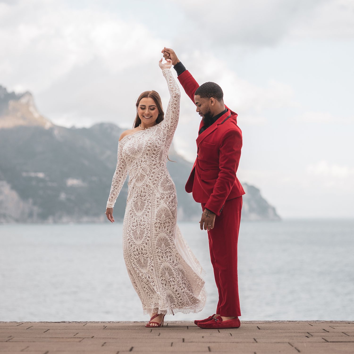 A couple dancing on an oceanfront boardwalk, with the bride in a lace dress and groom in a red suit against coastal cliffs.