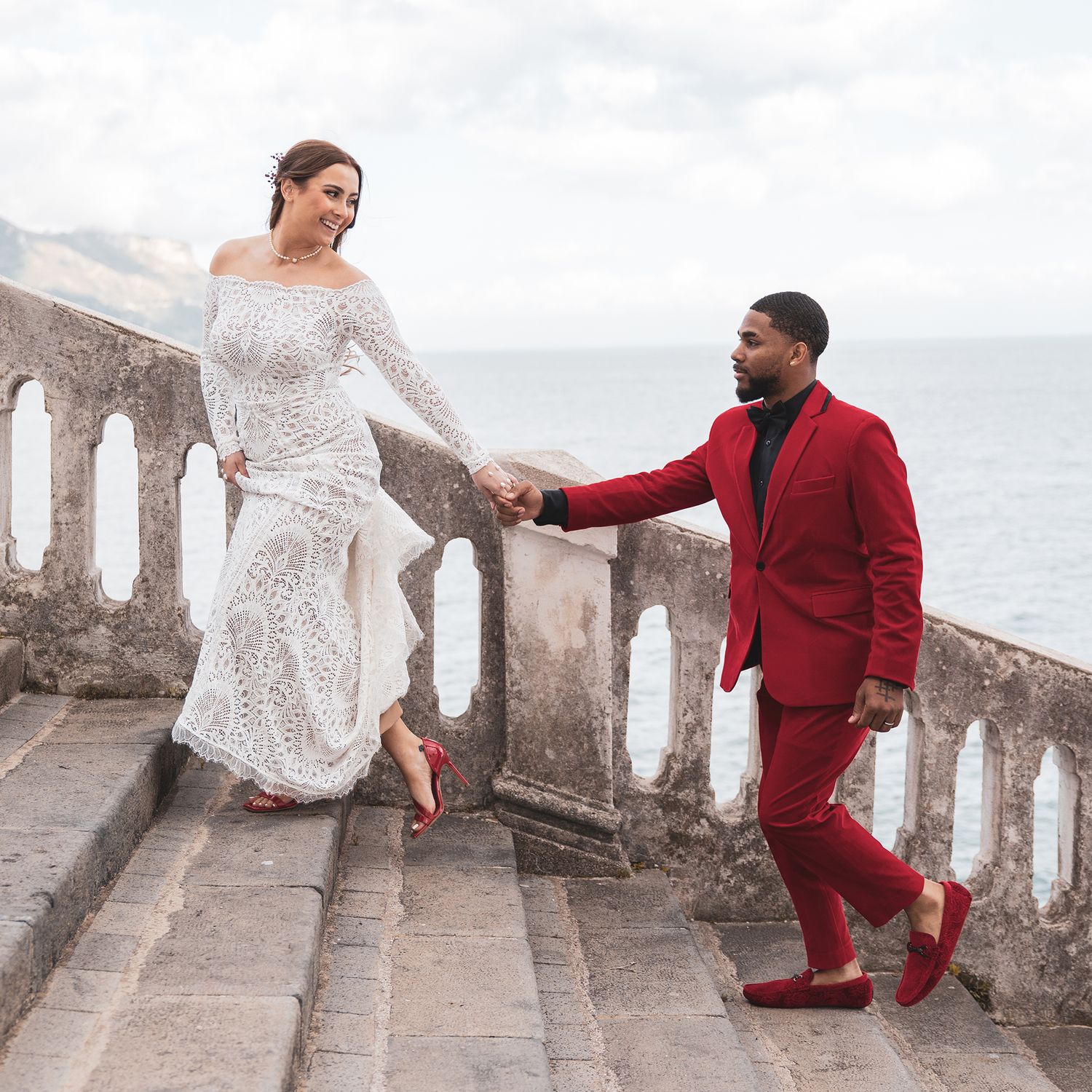 A couple in formal attire, with a white lace dress and red suit, pose together on stone stairs by the ocean.