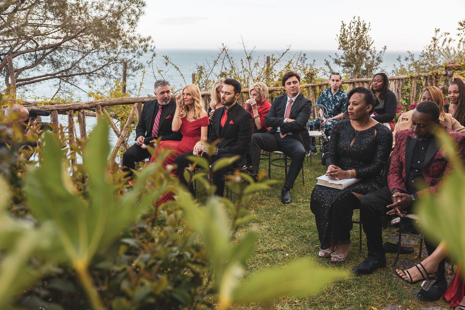 Group of people sitting outdoors on a hillside during an event with plants and foliage in the foreground.