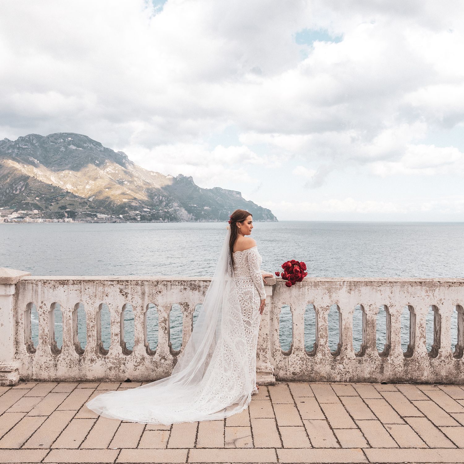 A bride in a flowing white gown and veil holds red flowers on an ornate balcony overlooking a coastal mountain landscape.