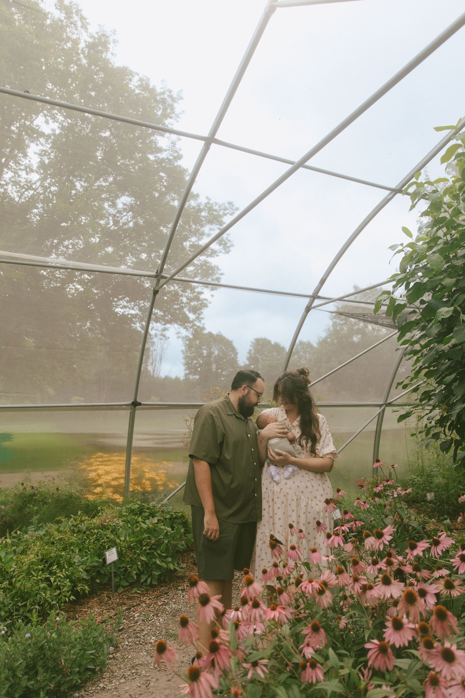 A couple with a newborn baby stands among pink echinacea flowers inside a misty greenhouse.