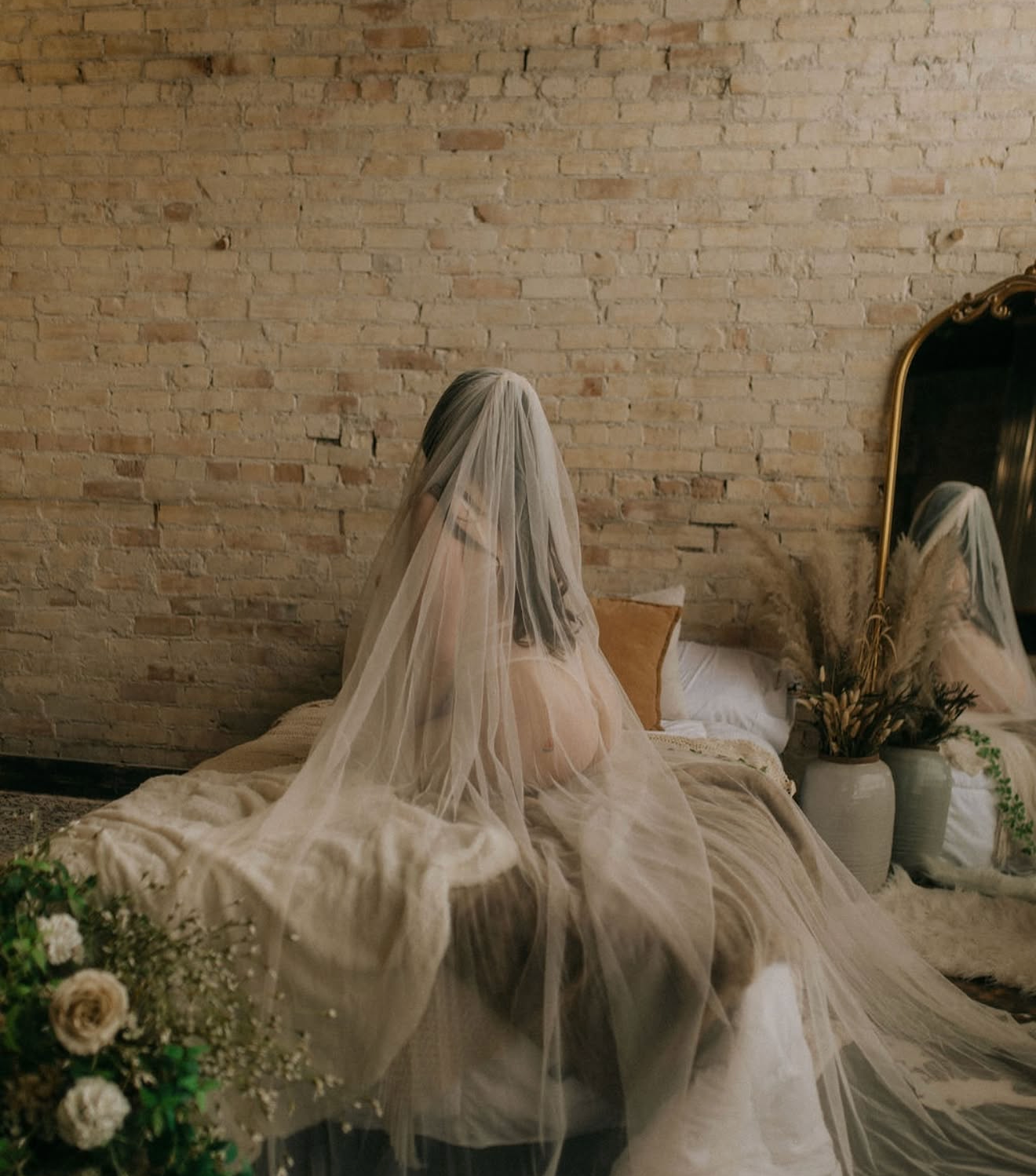 Bride in a flowing white veil sitting on a bed facing a brick wall, with floral arrangements nearby.