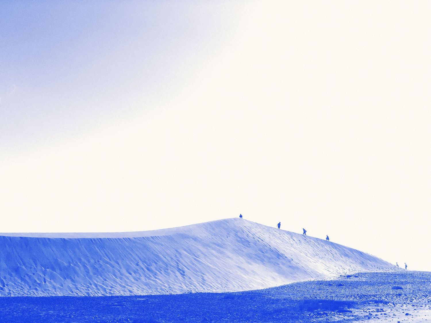 Blue-tinted desert dune landscape with tiny silhouettes of people walking along the ridge under a pale sky.