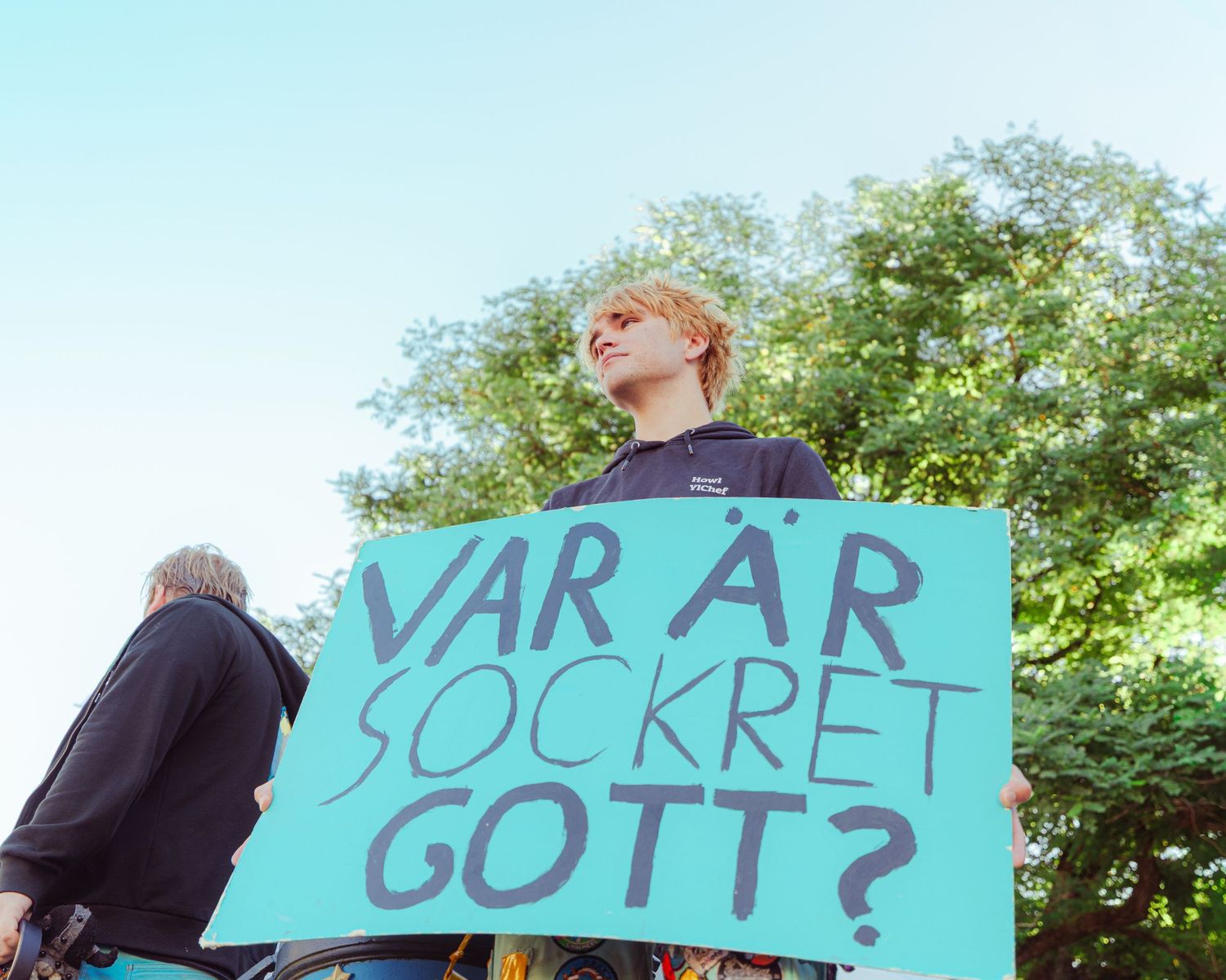 Person holding a teal sign reading VAR ÄR SOCKRET GOTT? at an outdoor protest with trees in background.