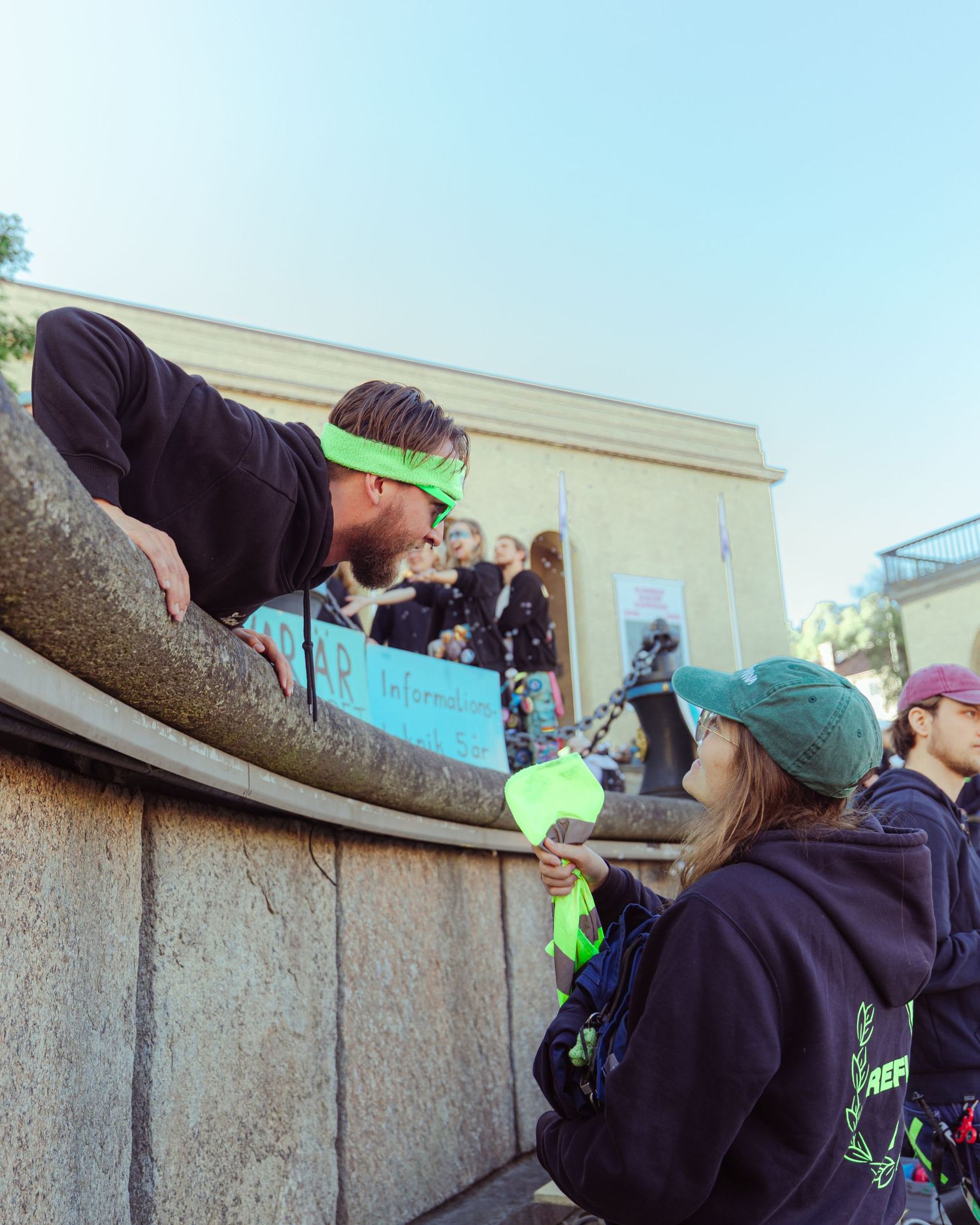 Man in green headband leans over stone wall talking to woman in green cap at outdoor event.