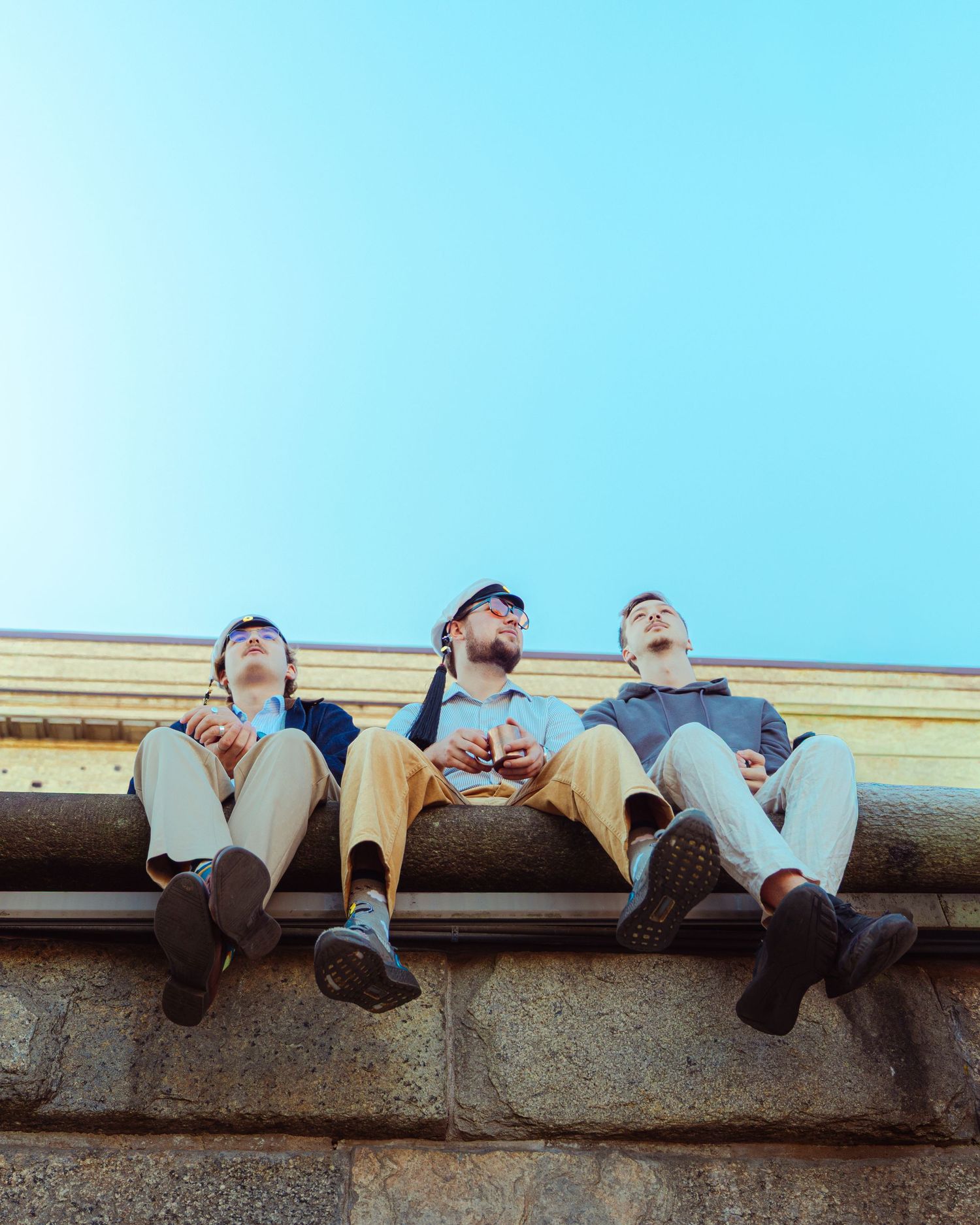 Three young men sitting on a stone ledge looking up at a clear blue sky outdoors.