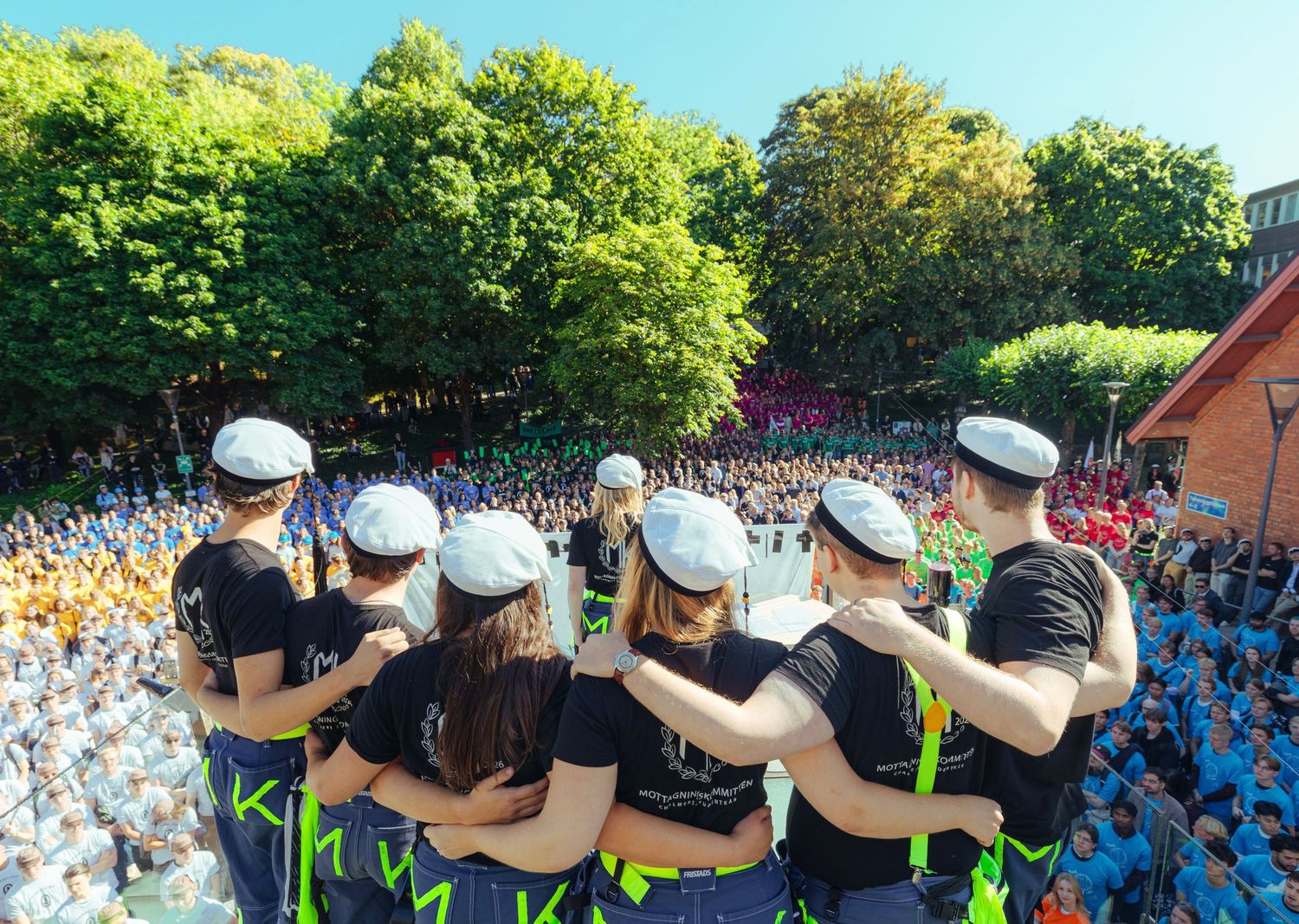 Group of students in black shirts and white caps stand together overlooking a large colorful crowd outdoors.