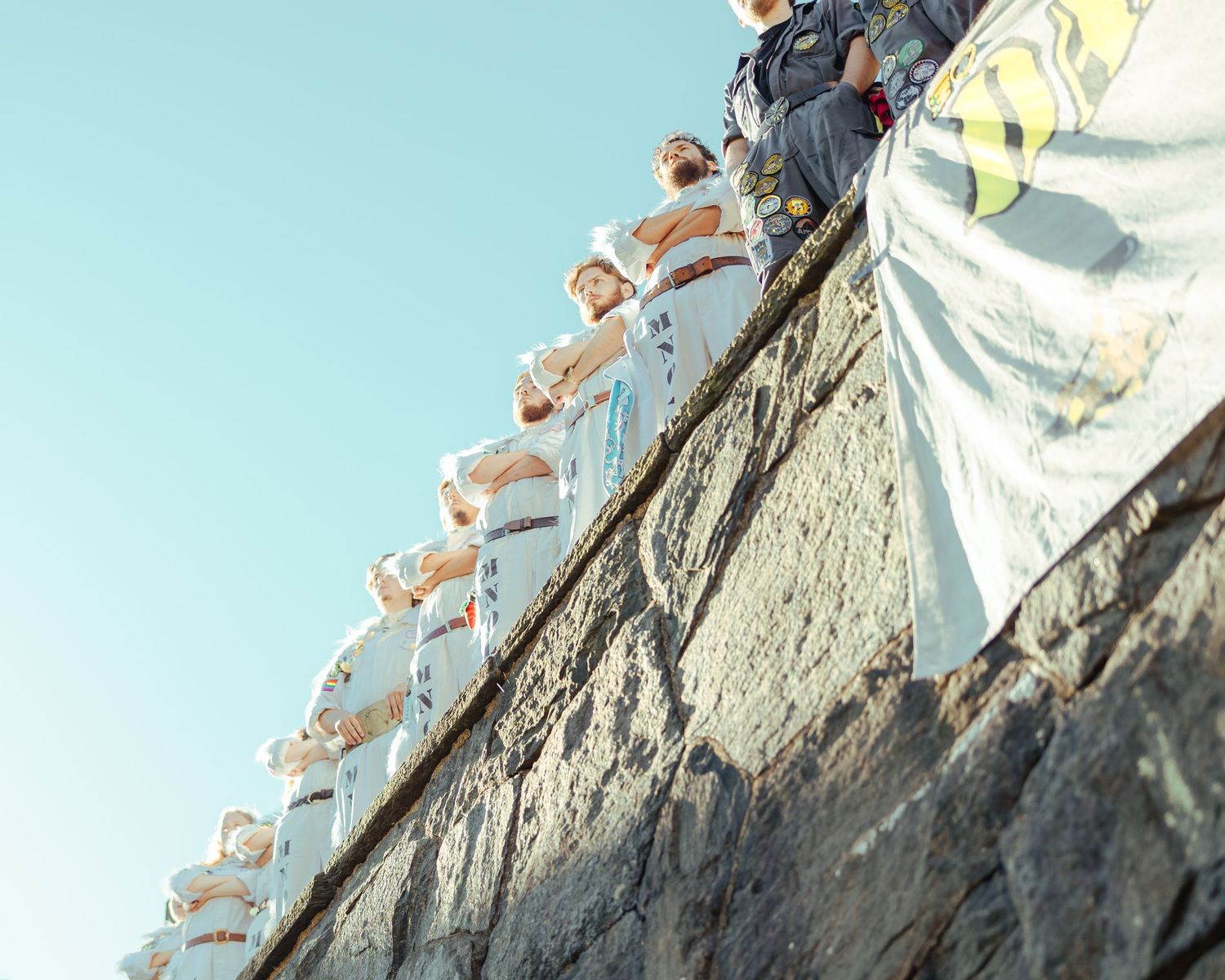 Group of people in white uniforms standing atop a stone wall against a clear blue sky.