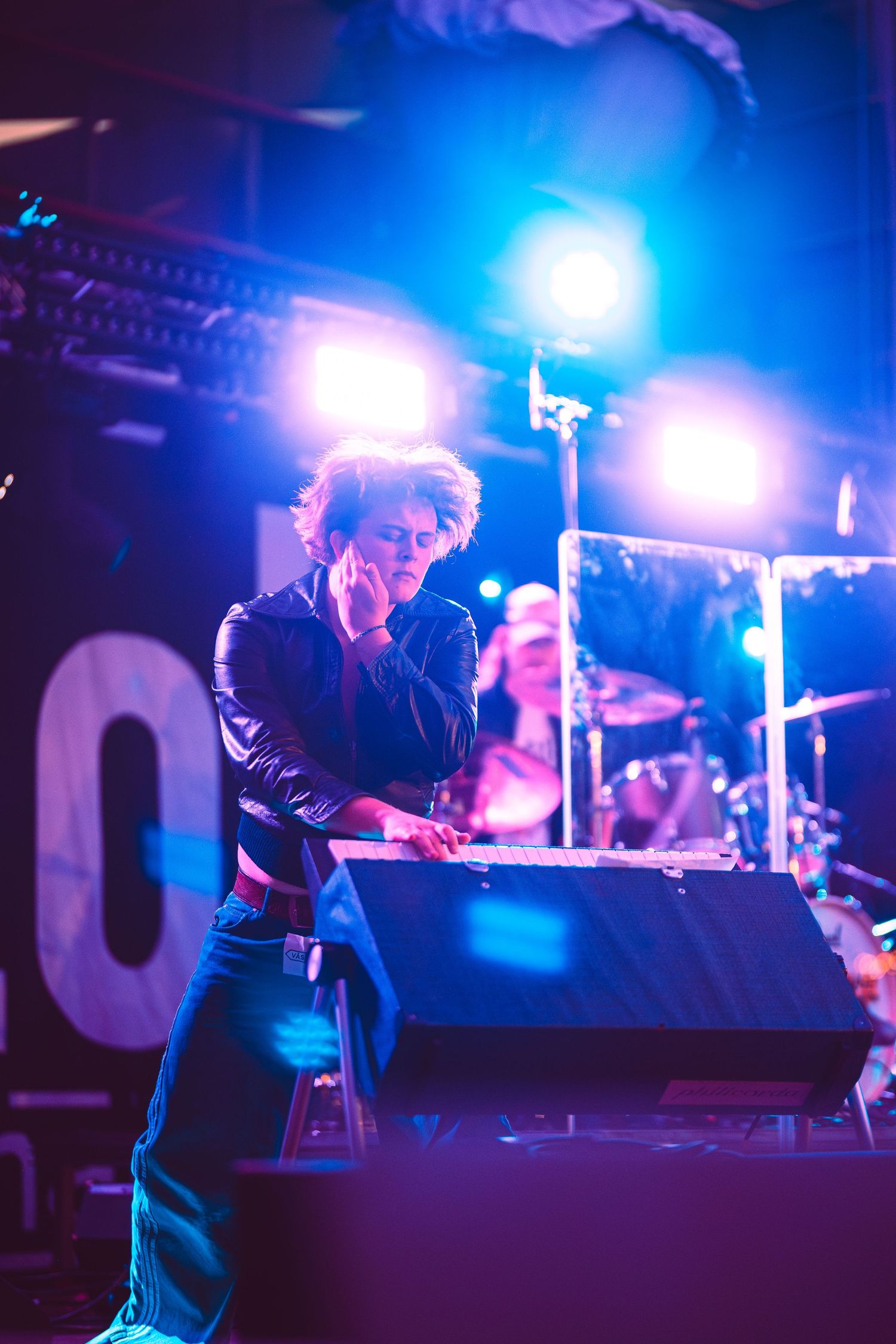 Performer leaning over stage monitor under blue and purple concert lights with band in background.