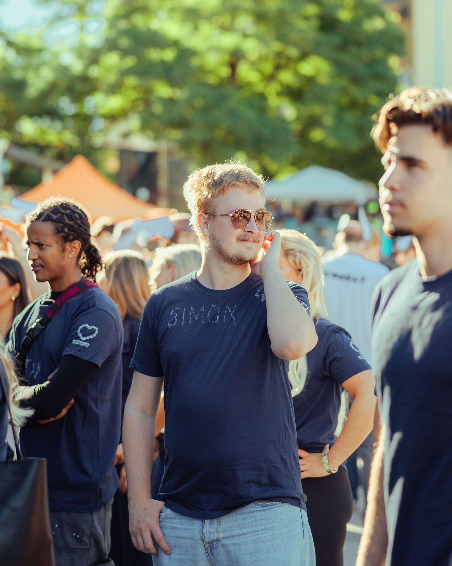 Young man in a navy Simon t-shirt and sunglasses stands at an outdoor festival crowd on a sunny day.