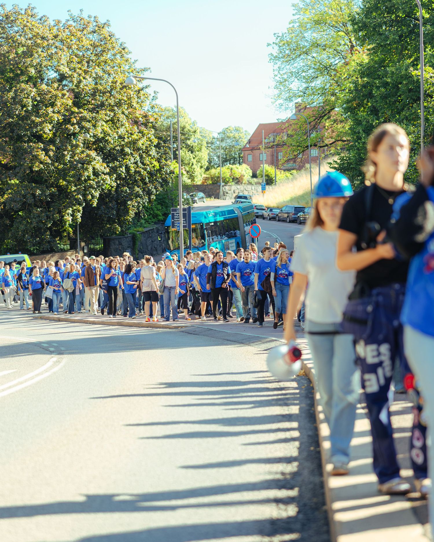 Large group of students in blue shirts marching along a sunny tree-lined street during a school event.
