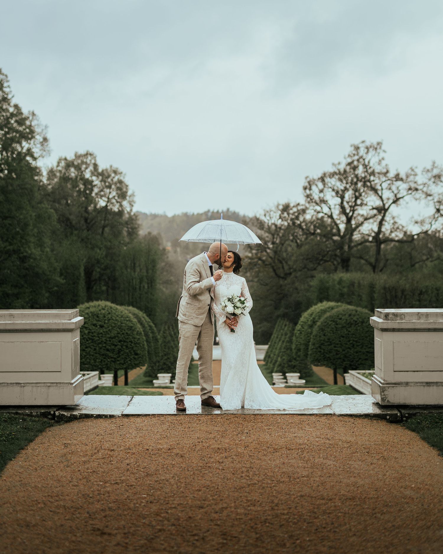 Wedding couple kissing under umbrella in elegant garden venue on a rainy day.
