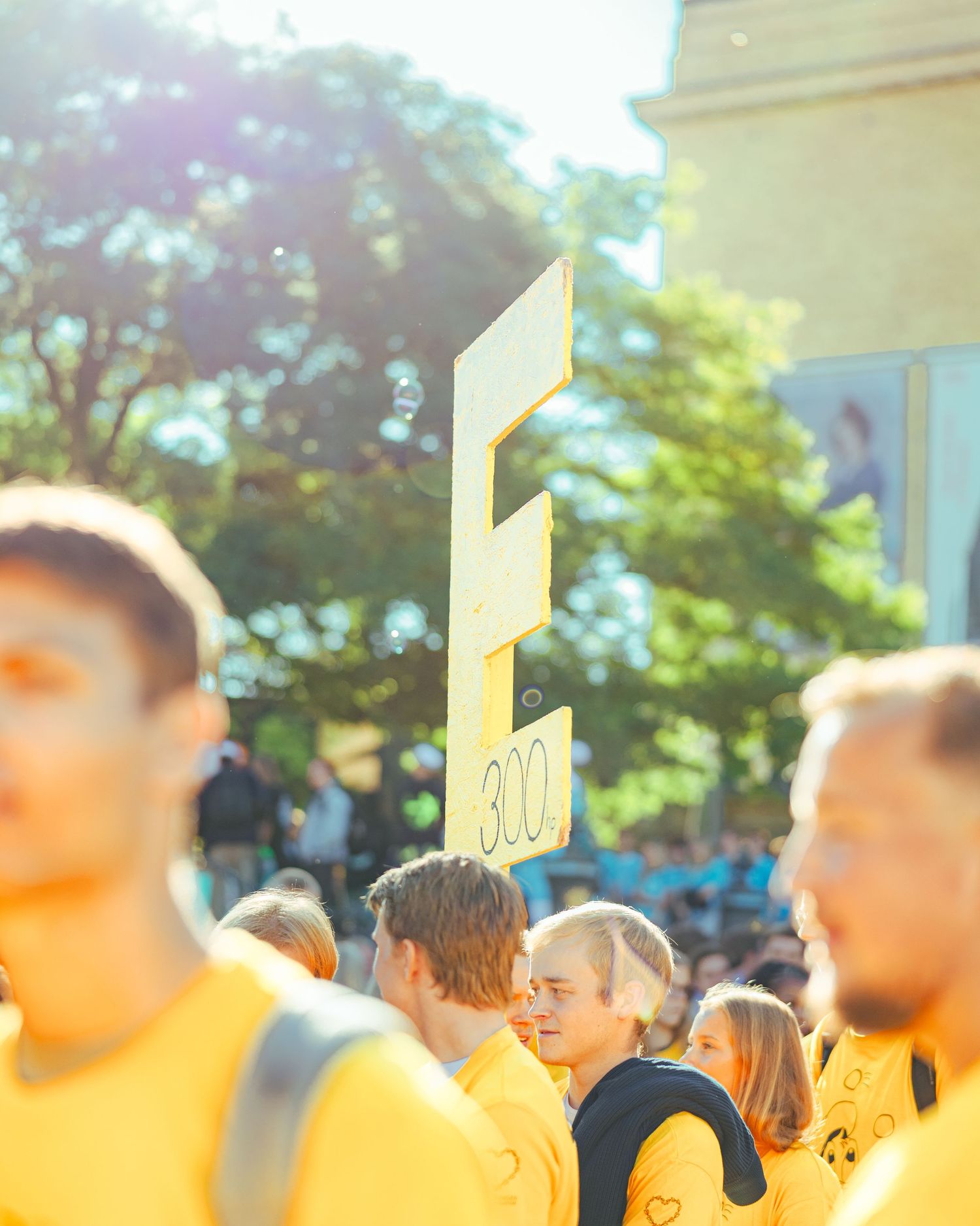 Crowd of people wearing yellow shirts at an outdoor event, holding a sign with the letter E and number 300.