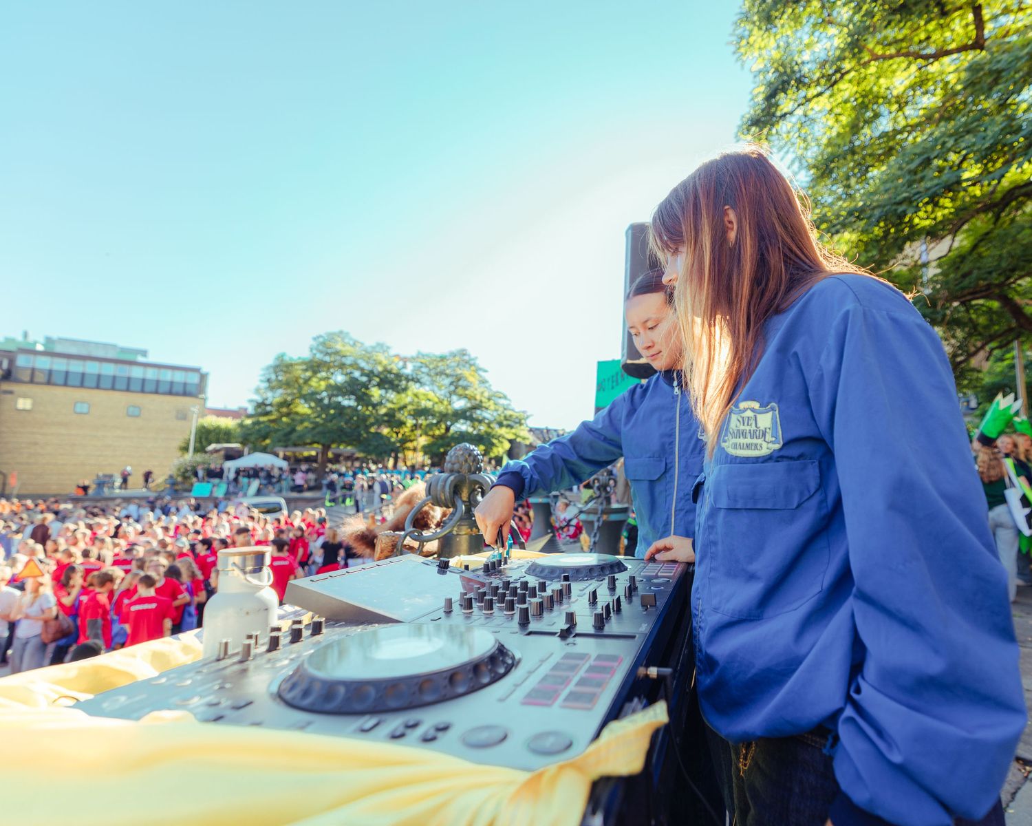 Two female DJs in blue jackets perform at an outdoor event with a large crowd gathered in the background.
