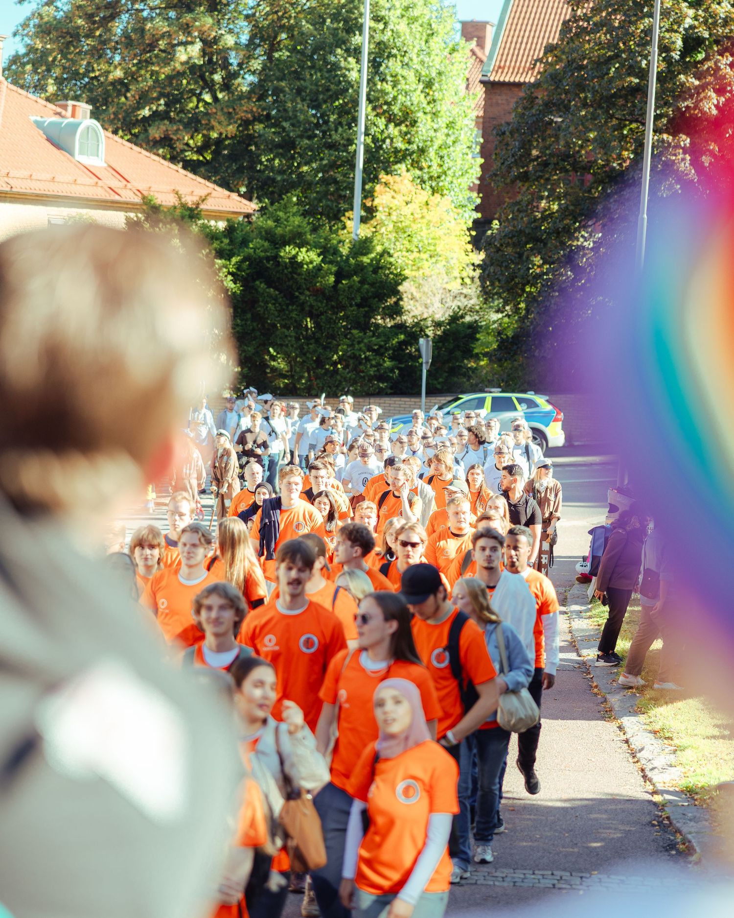Large group of people wearing orange shirts walking together outdoors on a sunny day near trees and buildings.