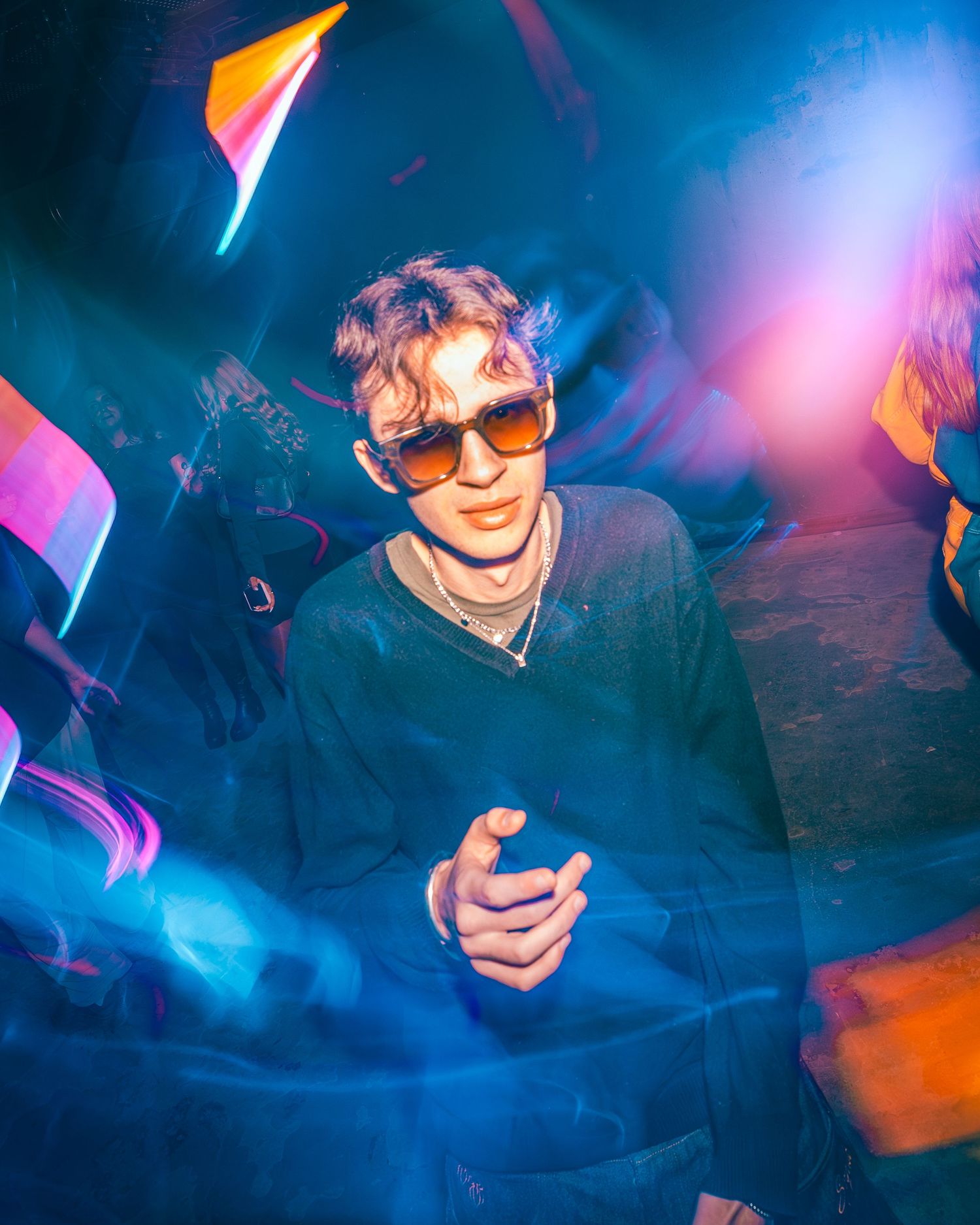 Young man in sunglasses surrounded by colorful neon lights at a vibrant nighttime event.