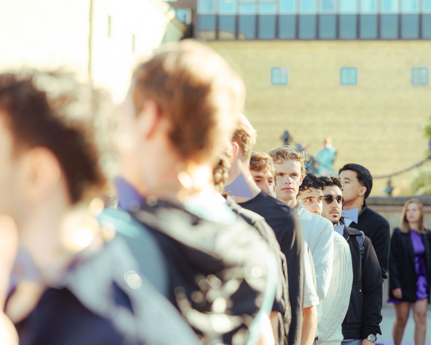 A group of young men standing in line outdoors near a building on a sunny day.