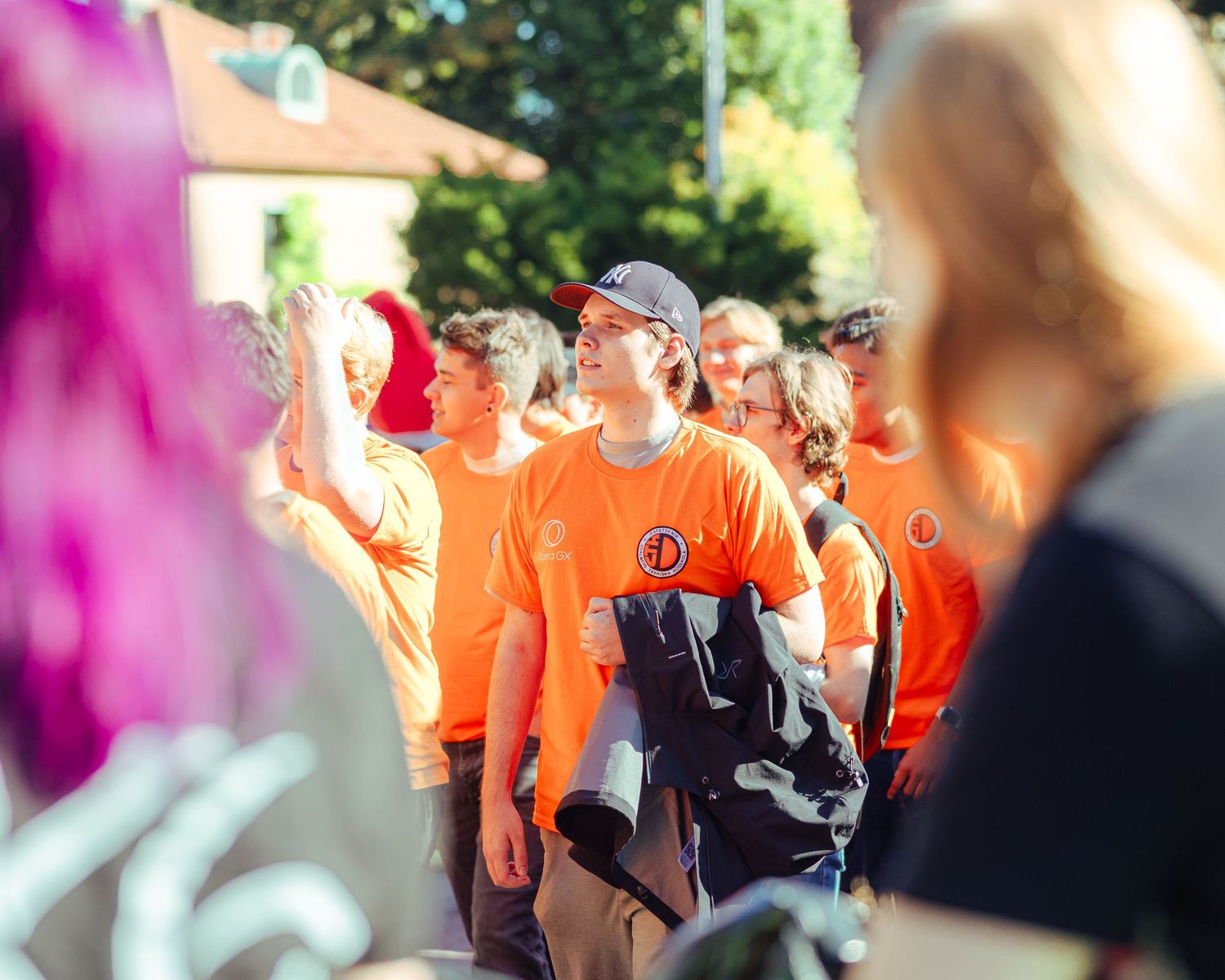 Group of people wearing matching orange t-shirts with a logo outdoors at a sunny community event.
