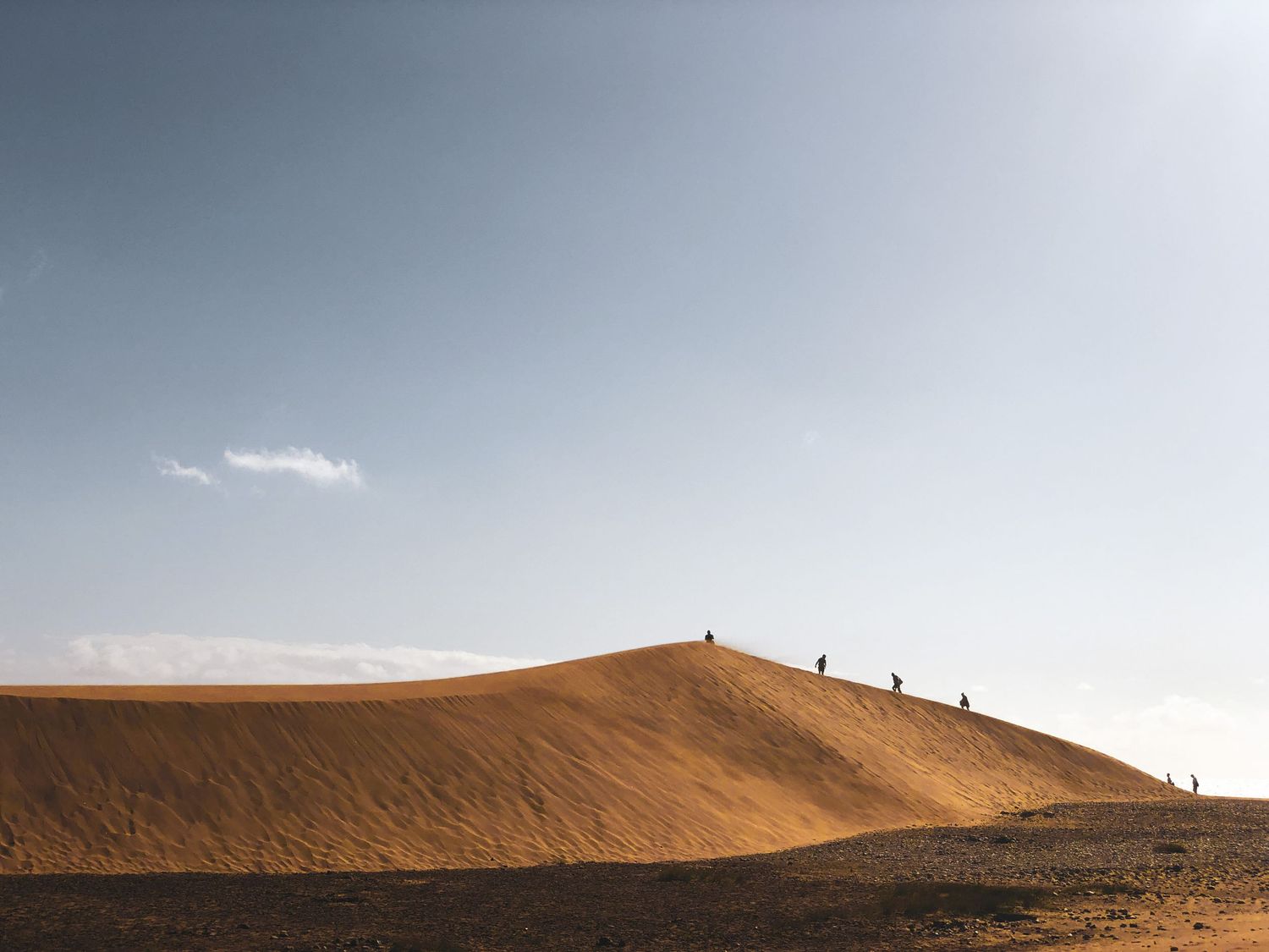 Tiny silhouettes of people climbing a vast golden sand dune under a clear sky.