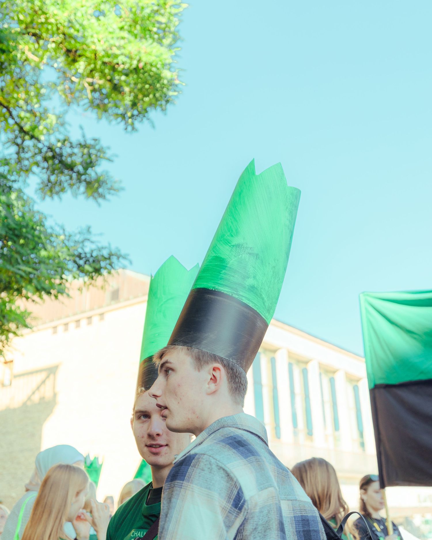 Man wearing a tall green and black paper crown at an outdoor rally with trees and a building in the background.