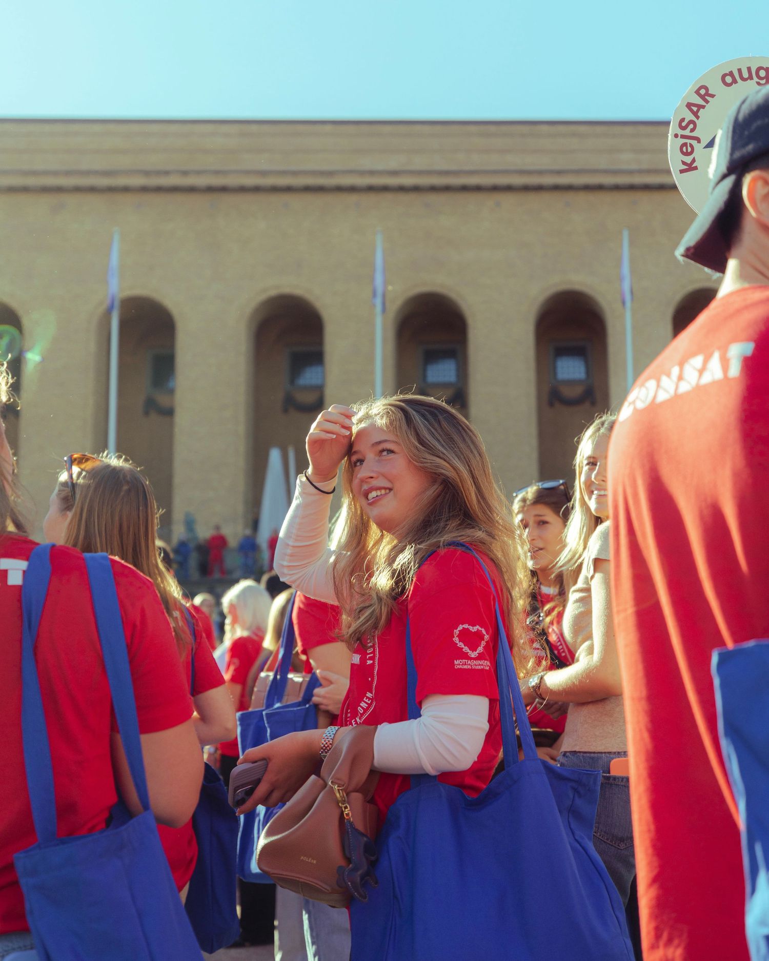 Smiling young woman in red vest and blue tote bag at outdoor event with crowd.