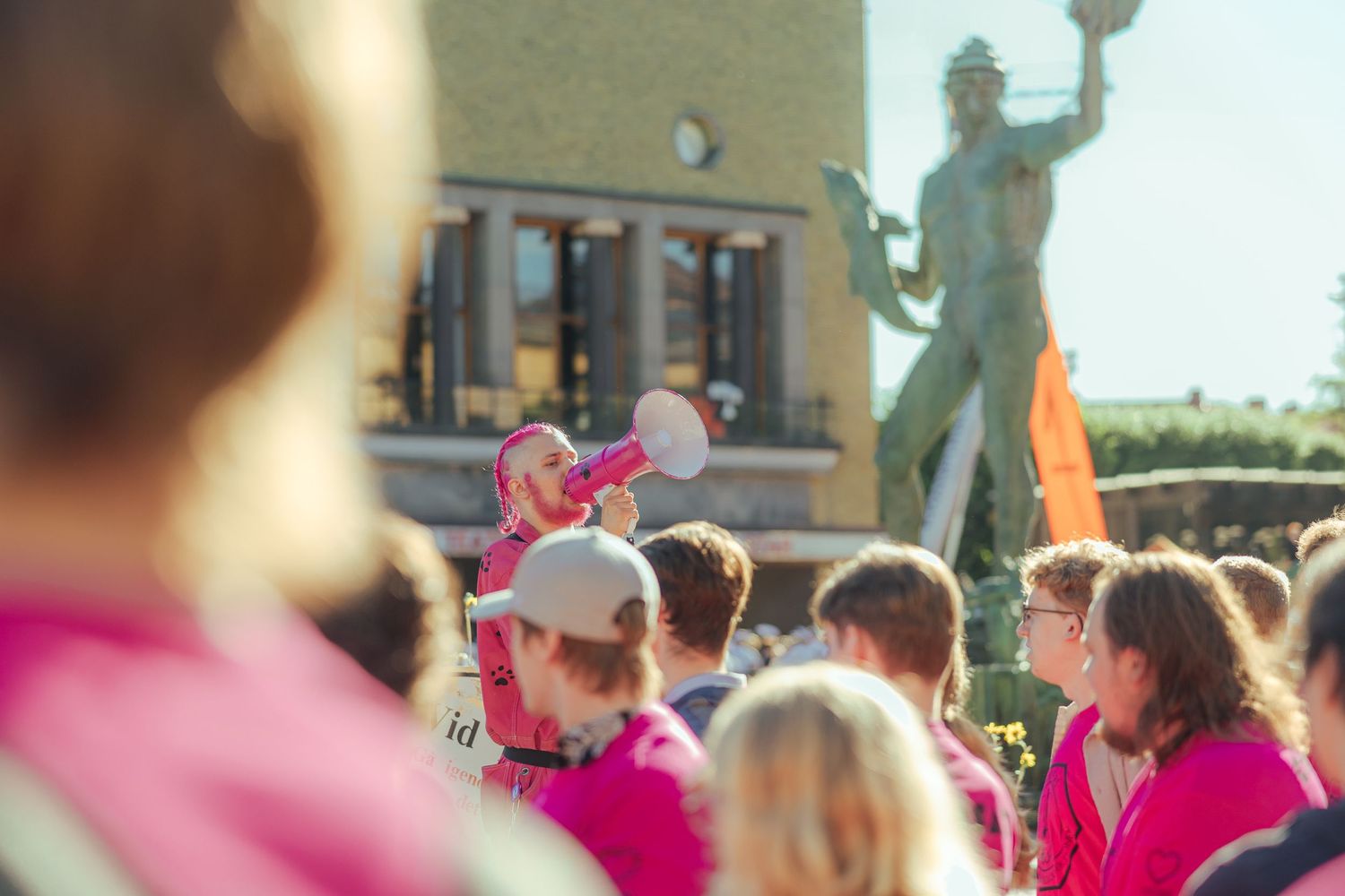Crowd in pink shirts gathered around a statue during an outdoor event on a sunny day.