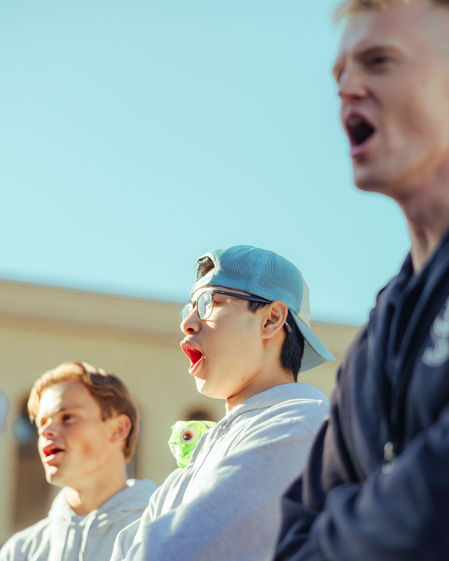 Three young men singing outdoors under a clear blue sky, one wearing a snapback cap and red lipstick.