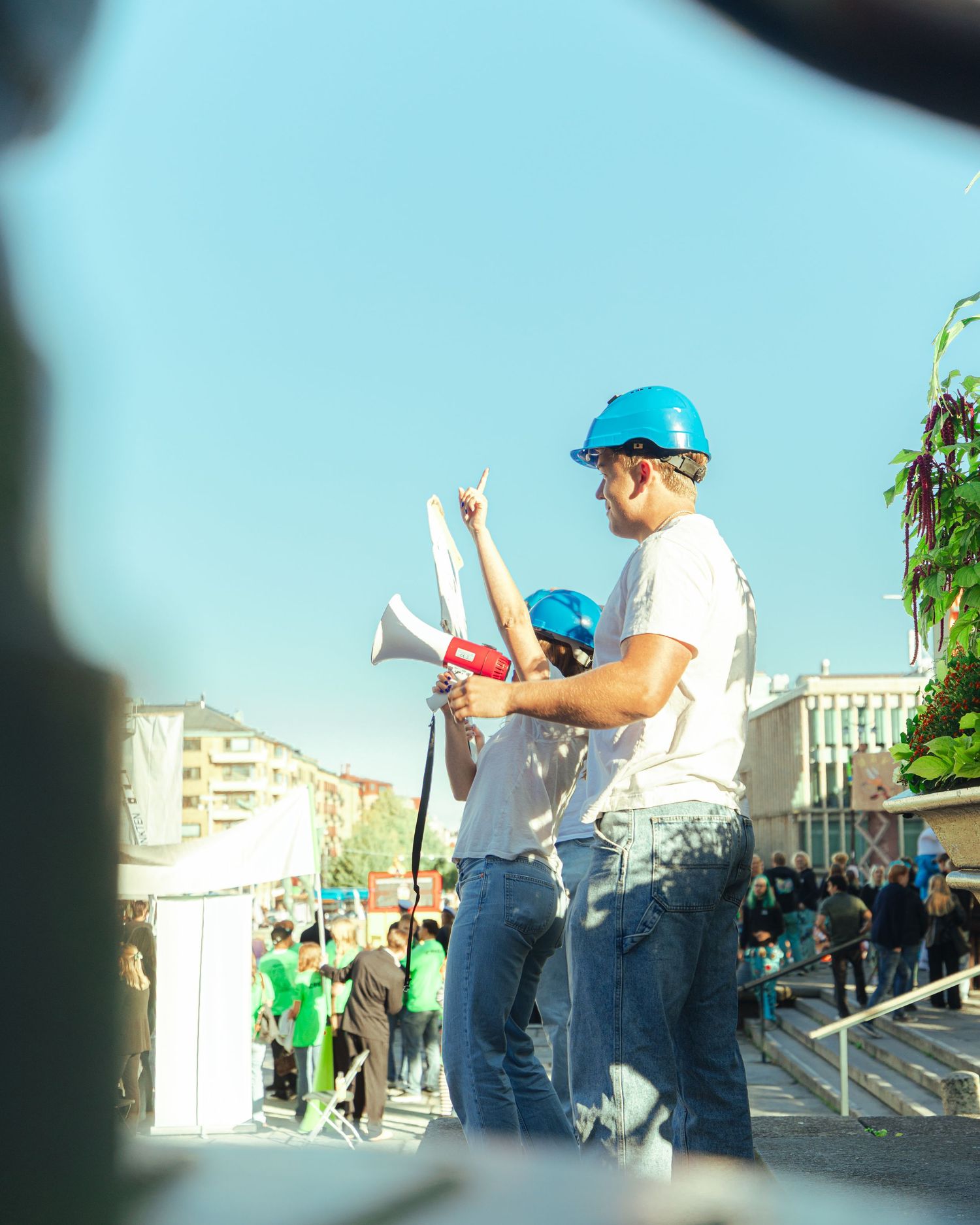 Two people wearing blue hard hats at an outdoor rally, one holding a megaphone and gesturing.