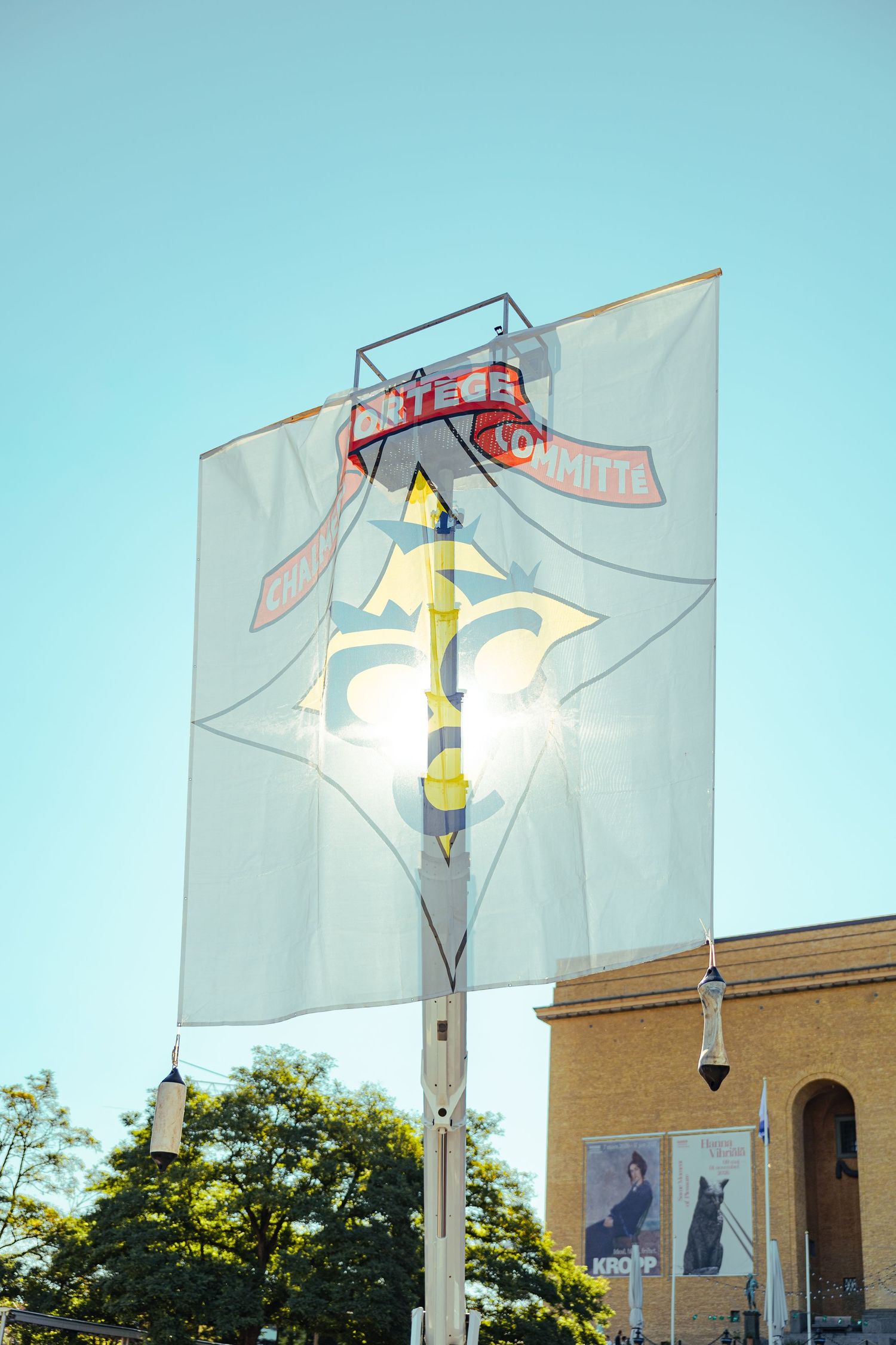 A large white banner with a fleur-de-lis design on a pole, backlit by sunlight against a clear blue sky.