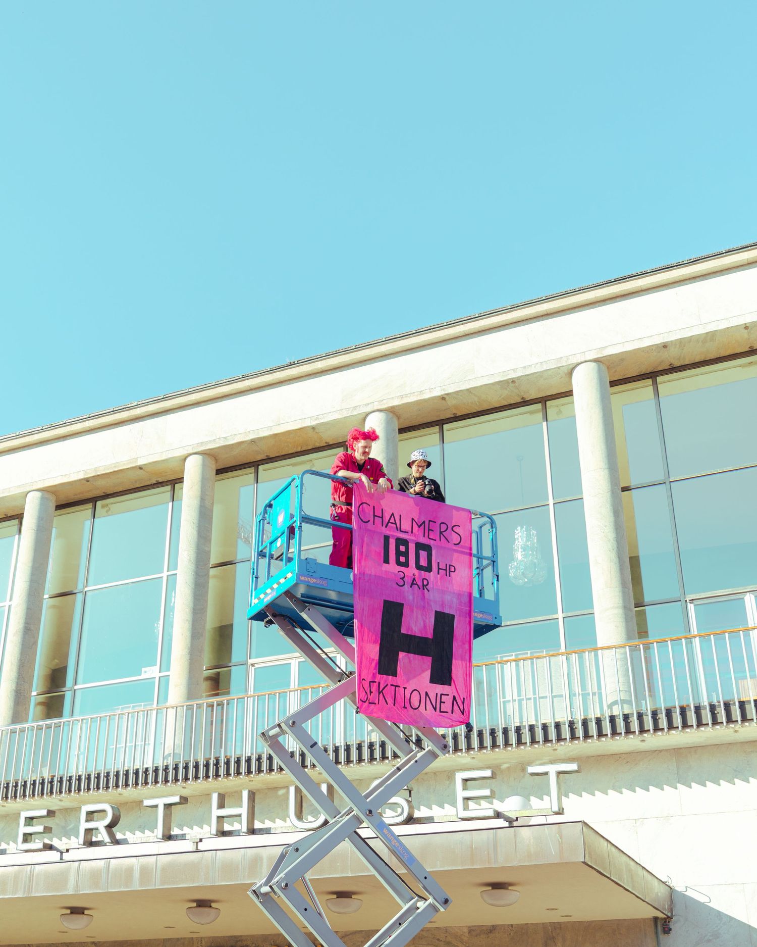 Two people on a cherry picker lift holding a pink periodic table element banner on a building facade.