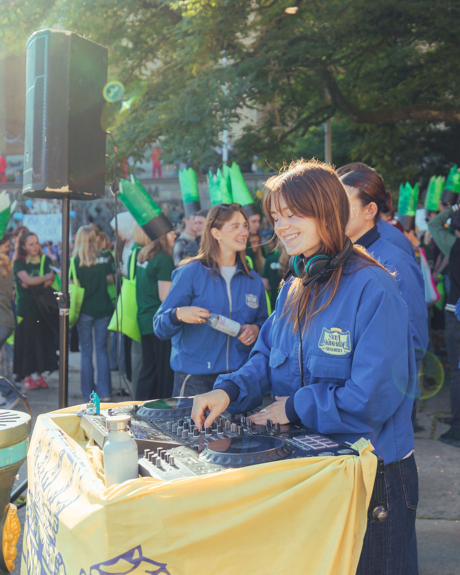 A smiling female DJ with headphones works a mixer at an outdoor event with a crowd waving green flags.
