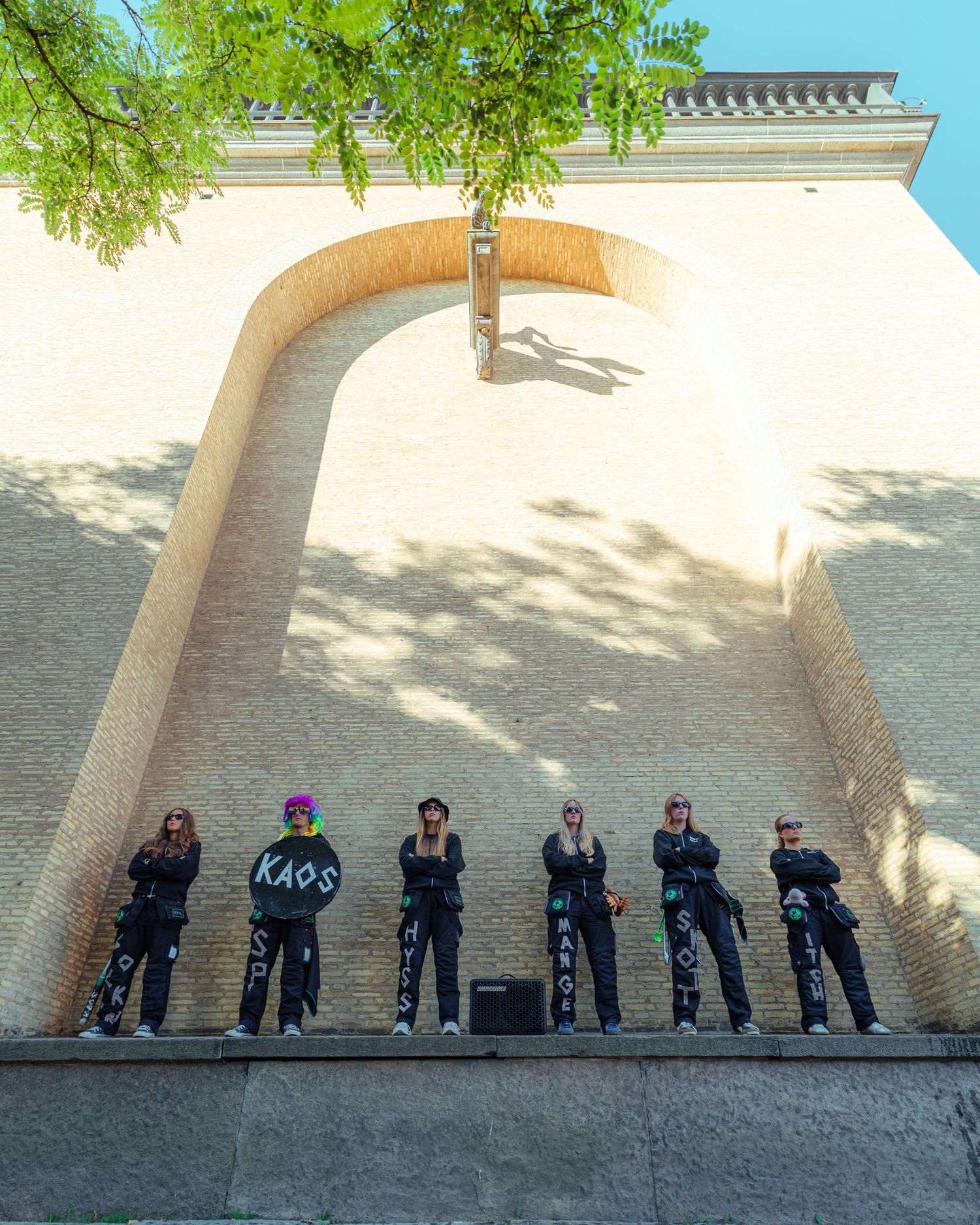 Group of seven people in matching black KAOS outfits posing against a curved brick wall outdoors.