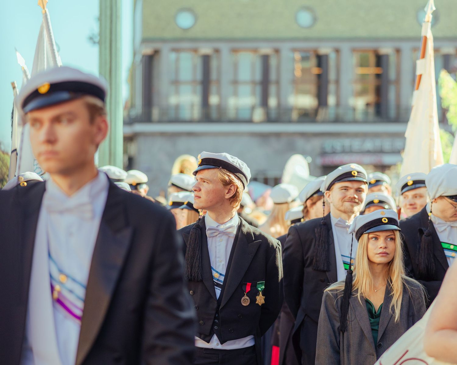Students in white caps and formal attire march in a ceremonial graduation parade in a European city square.