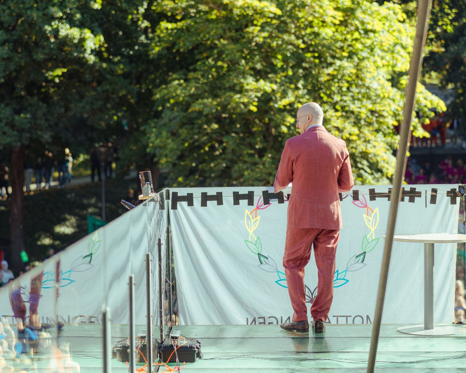 Man in red suit stands on outdoor stage before white banner, surrounded by lush green trees on sunny day.