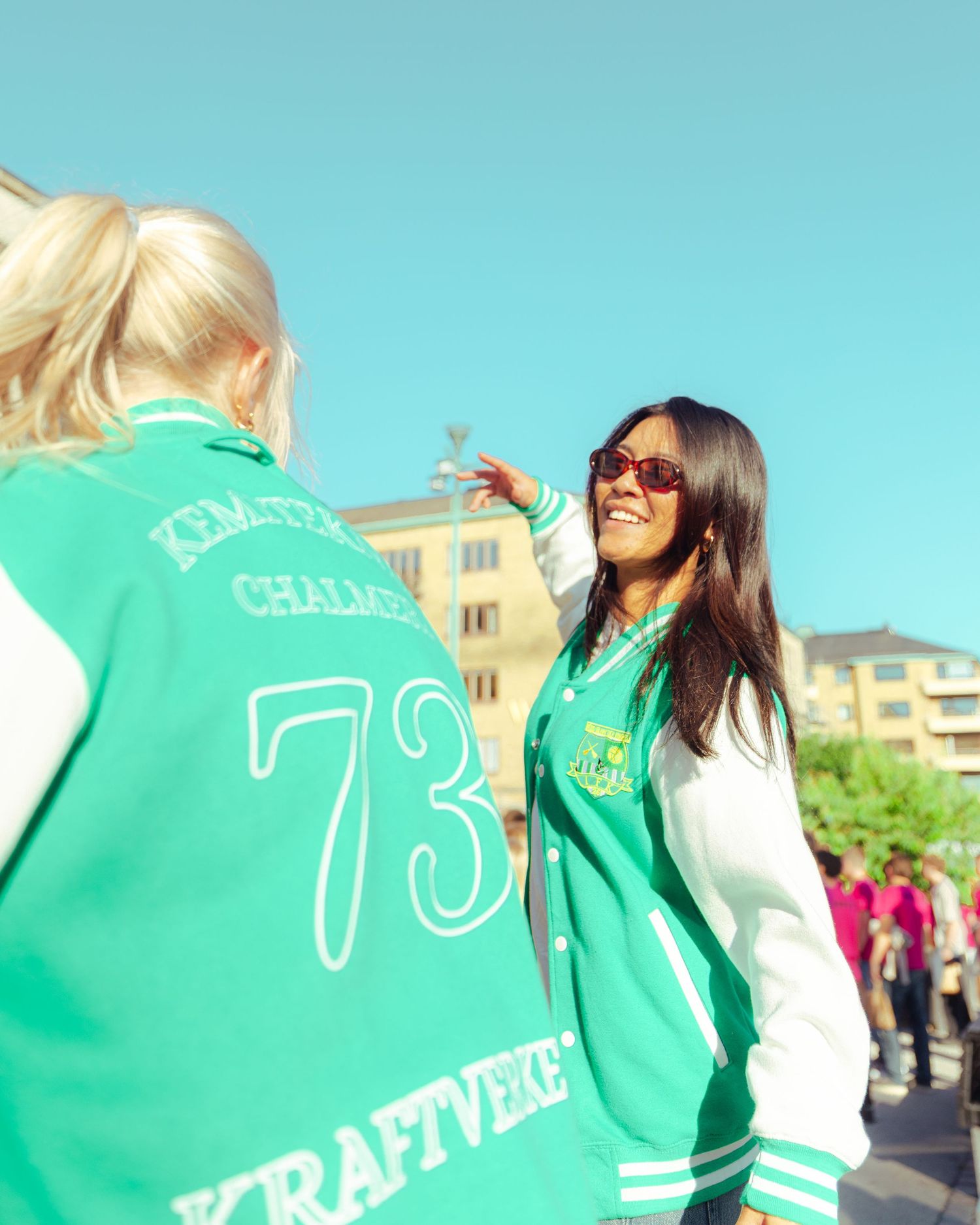 Two women wearing green and white varsity jackets with number 73 outdoors on a sunny day.