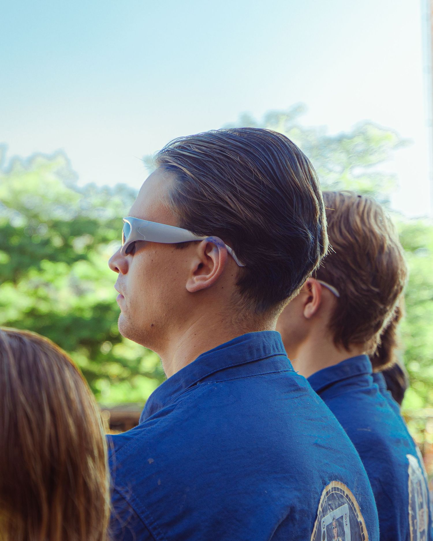 Group of people in blue uniforms with one wearing futuristic sunglasses outdoors with green trees in background.